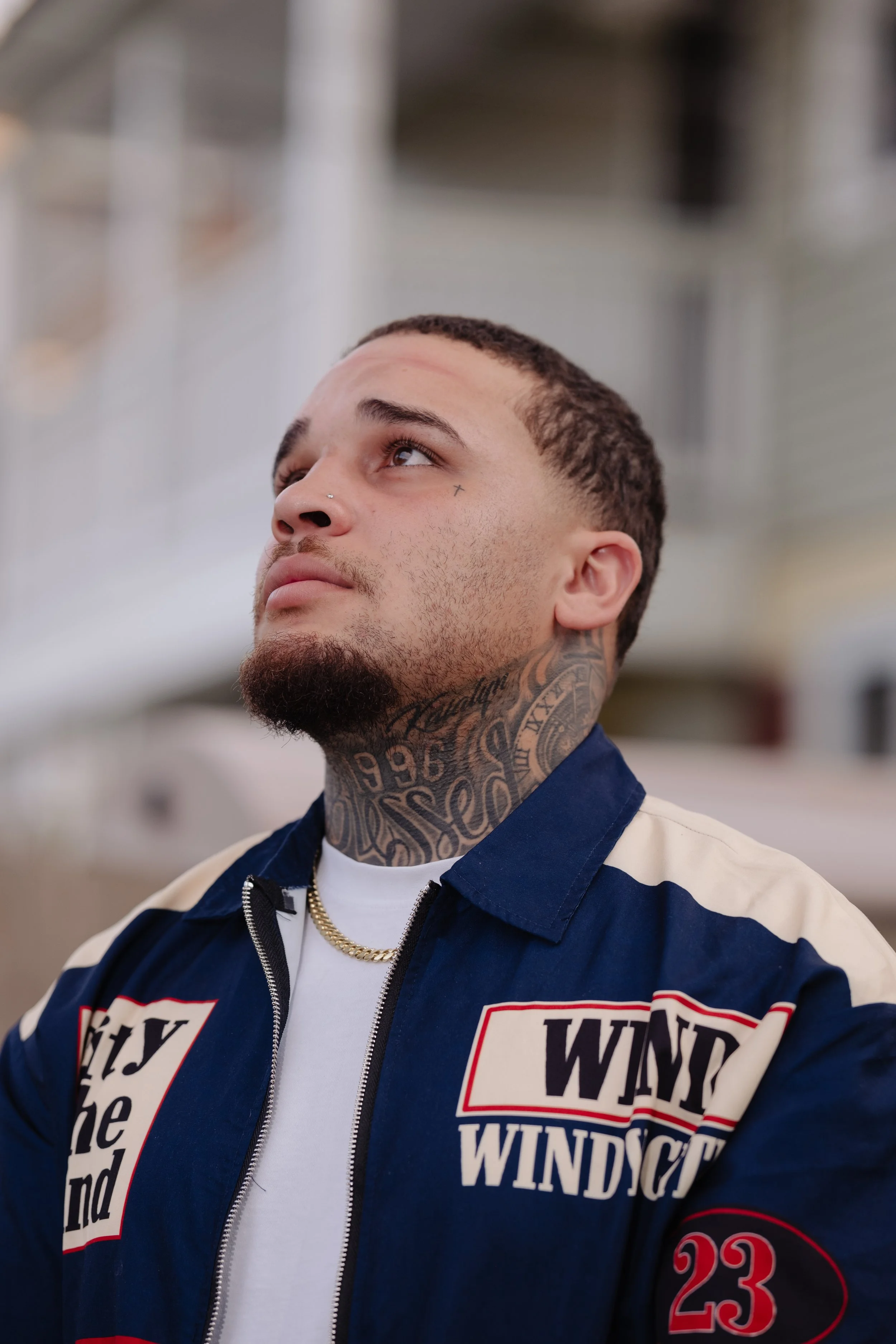 Close-up of a man with tattoos on his neck and face, looking upwards. He has short dark hair, a beard, a nose piercing, and wears a white t-shirt with a blue, red, and white jacket with patches and text. The background is blurred.