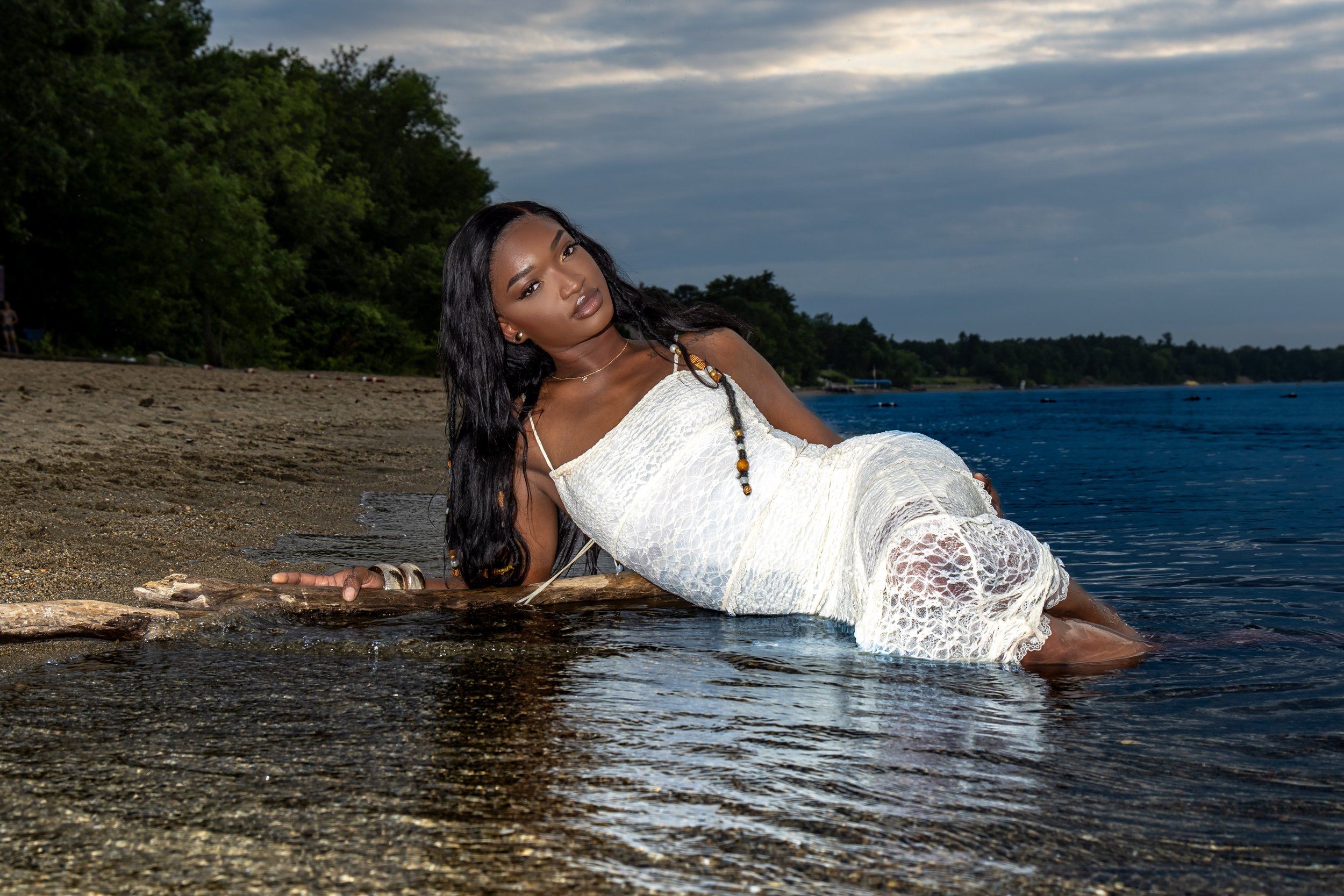 A woman with dark hair and jewelry, wearing a white dress, is lying on a log at the edge of a lake or river, with trees and cloudy sky in the background.