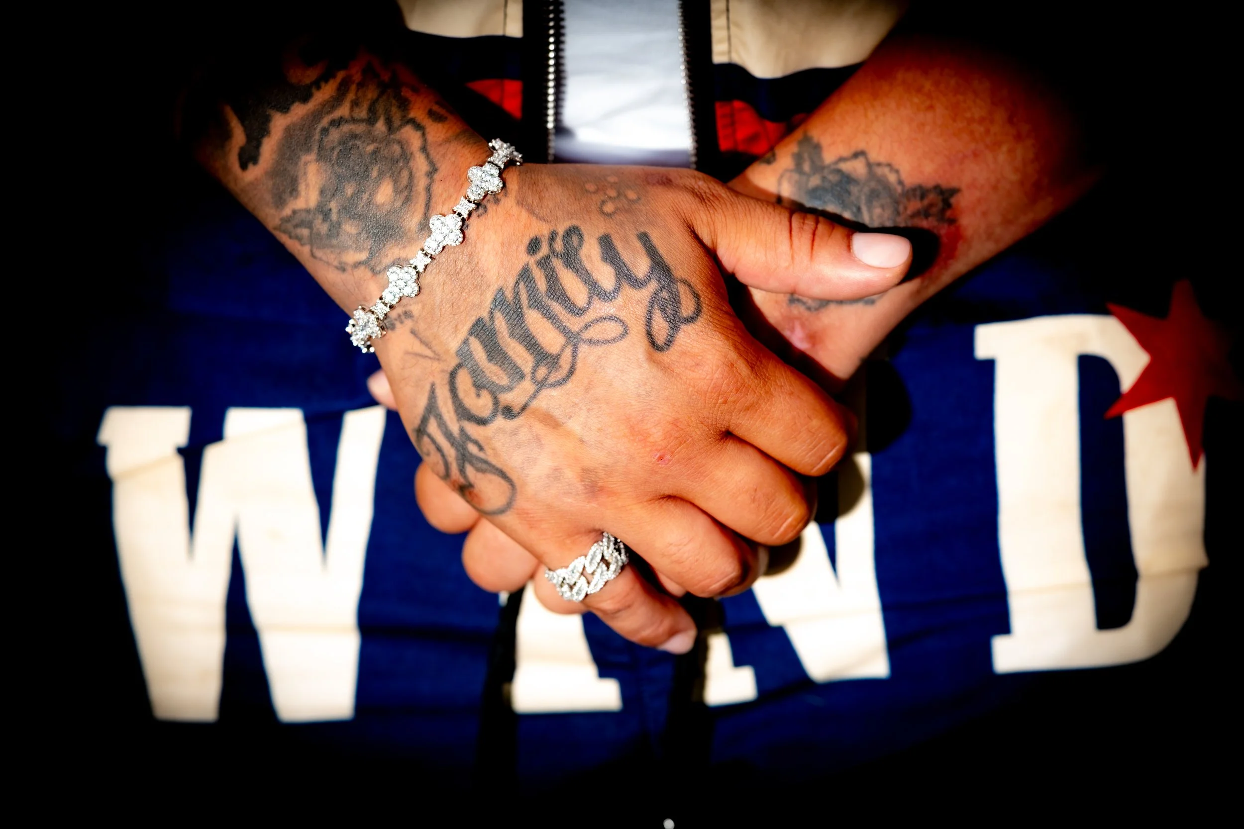 Close-up of two hands clasped together, showing tattoos and jewelry. One hand has a diamond bracelet and a large ring, with tattoos on the wrist and fingers. The person is wearing a dark shirt with a red and white star graphic.