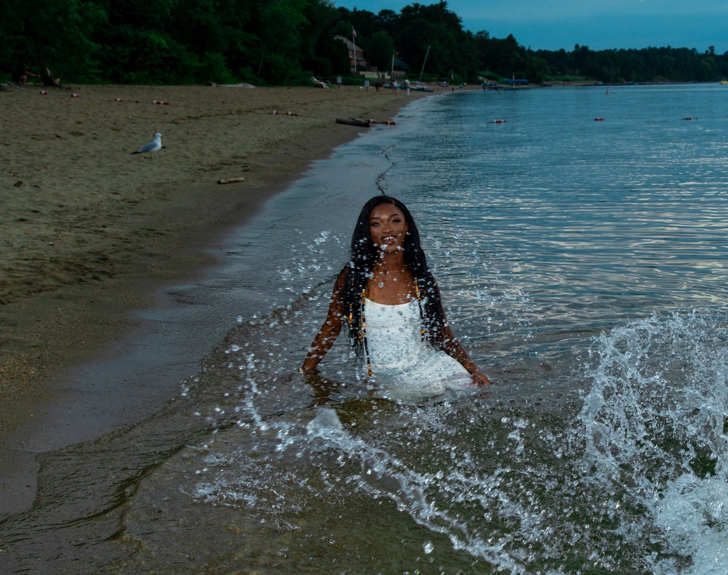 A woman with braided hair emerging from the water at a beach with a sandy shoreline, seagulls, and boats docked along a wooded hillside in the background.