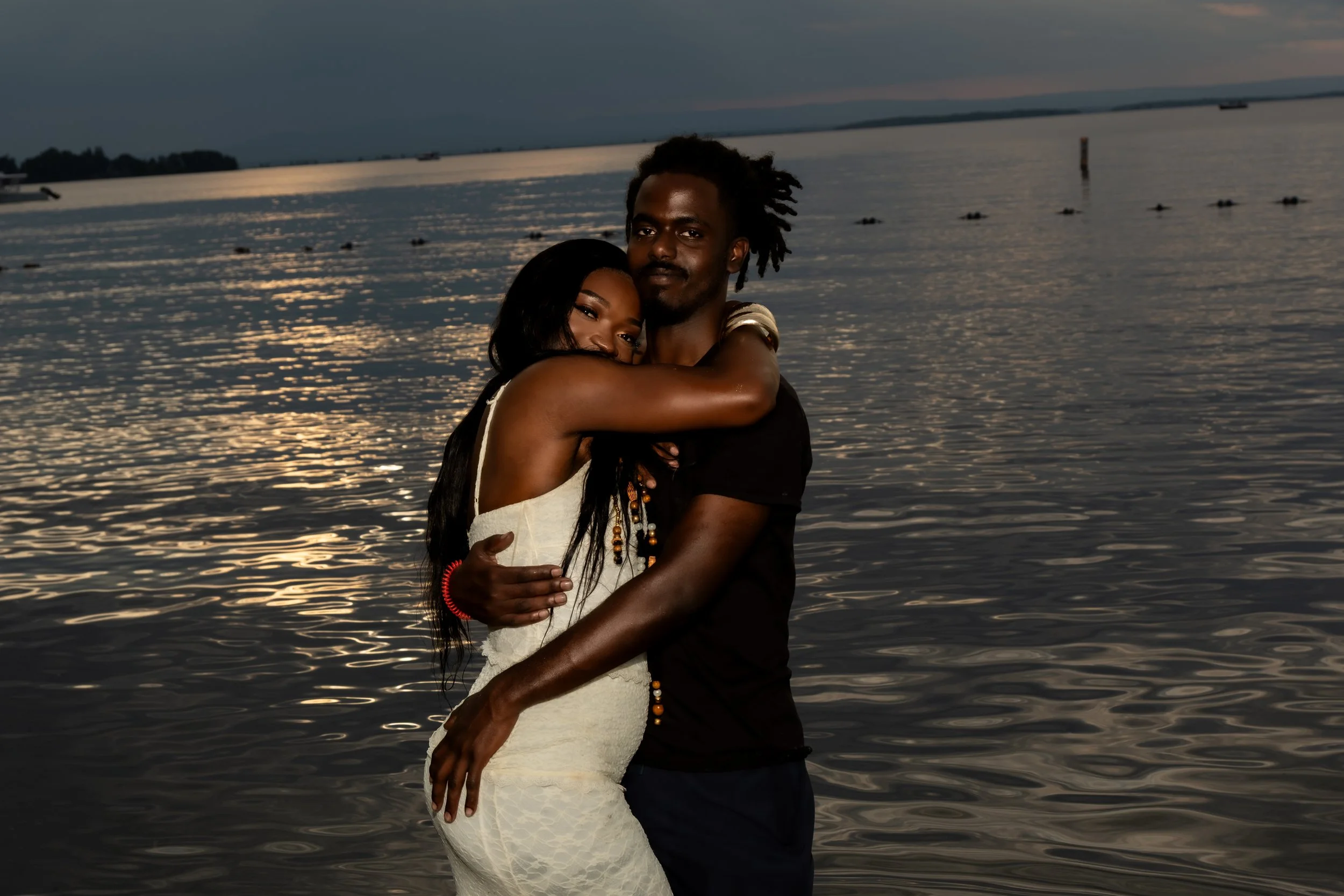 A couple embracing by a body of water during sunset, with the woman wearing a white dress and the man in a dark shirt.