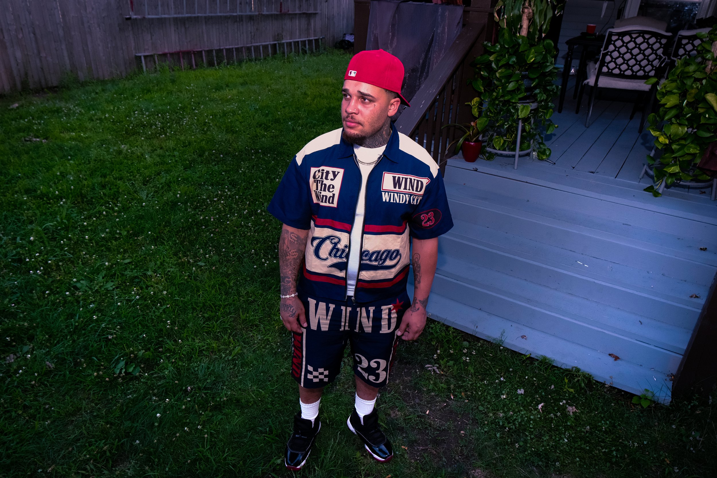 A young man in a Chicago-themed outfit, wearing a red cap backwards, standing outdoors on a grassy area near a wooden deck with chairs and plants, looking to his right.
