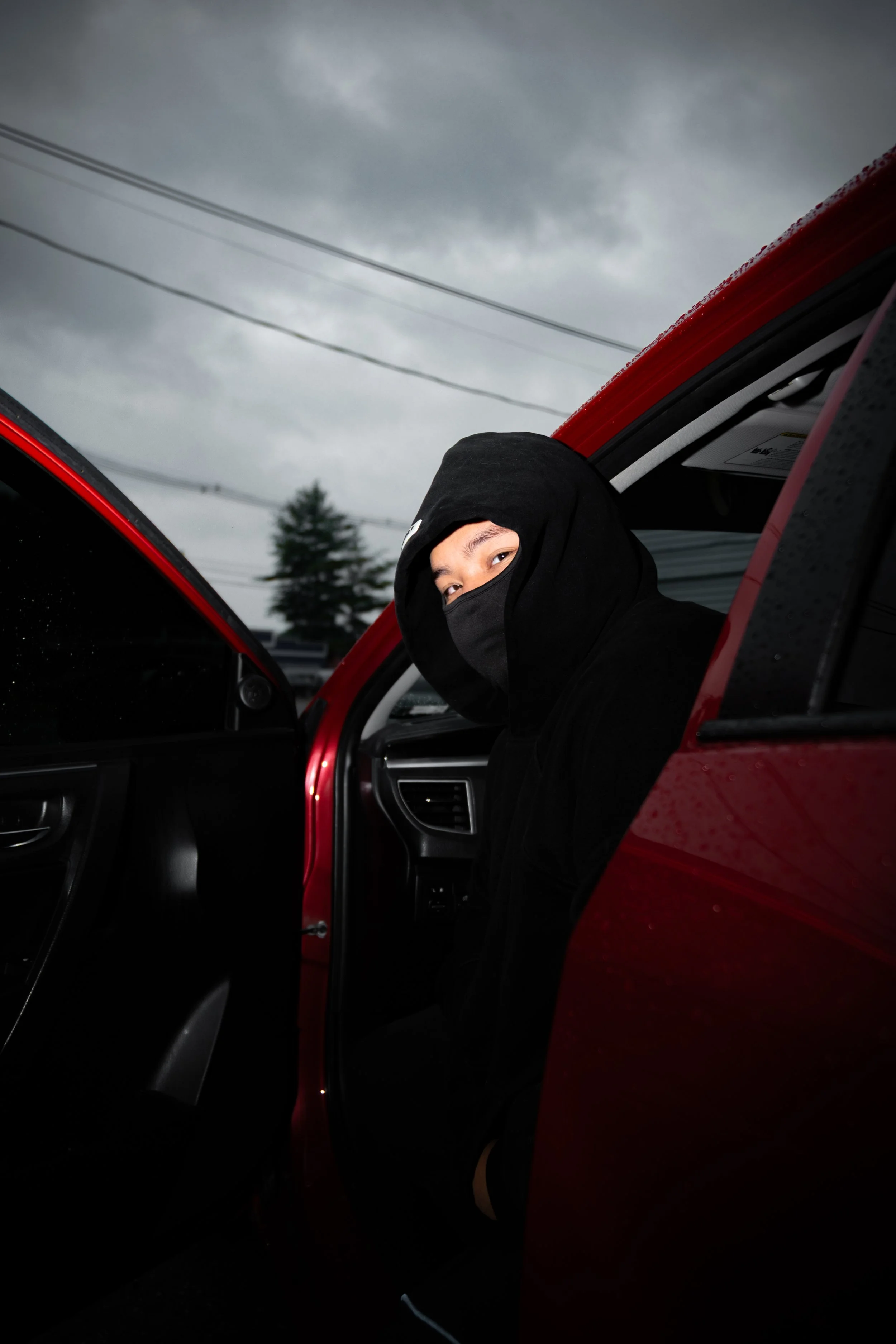 A person with a black hoodie and face mask sitting in a red car with the door open on a cloudy day.