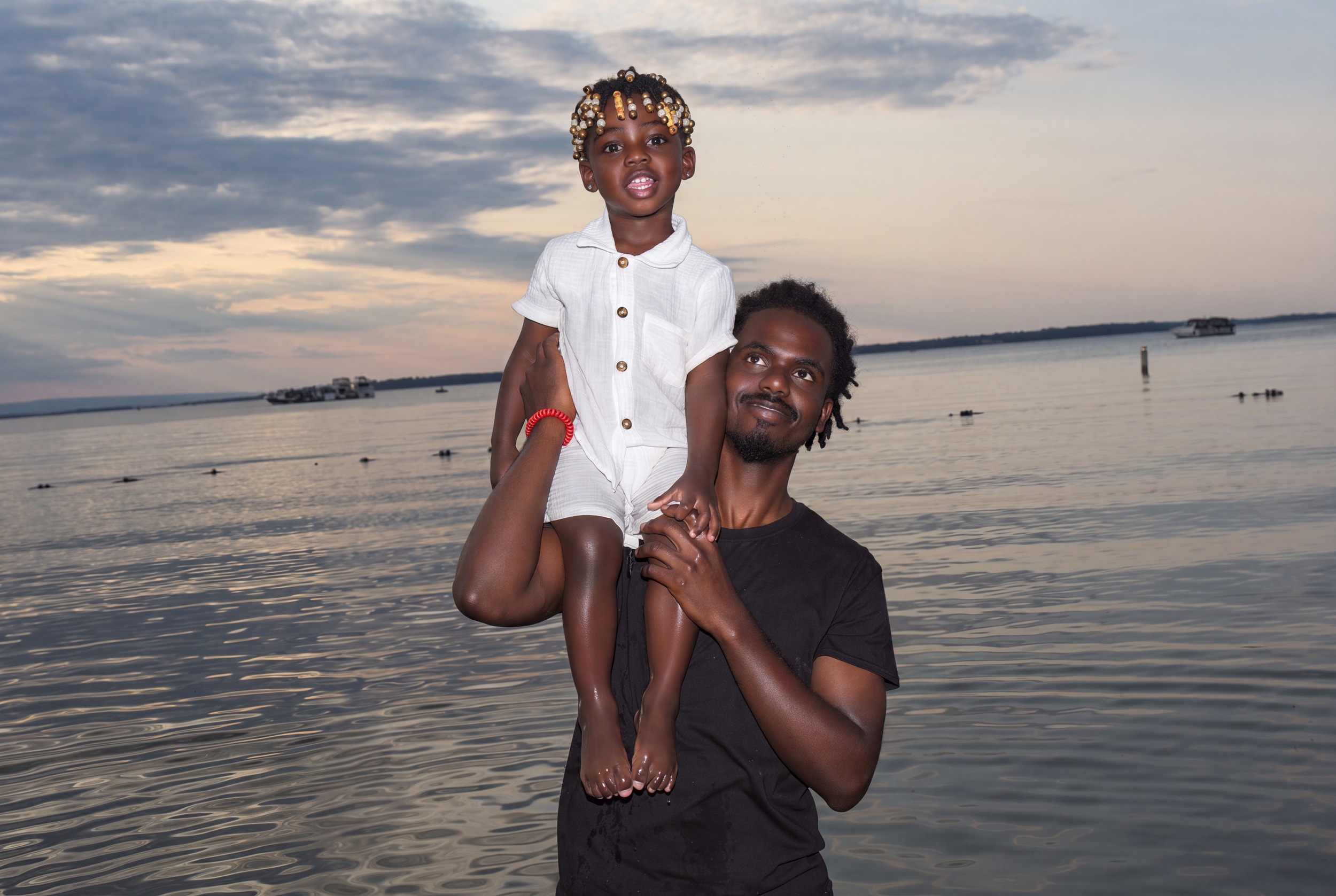 A man holding a young girl on his shoulder near a body of water, with boats in the distance and a cloudy sky in the background.