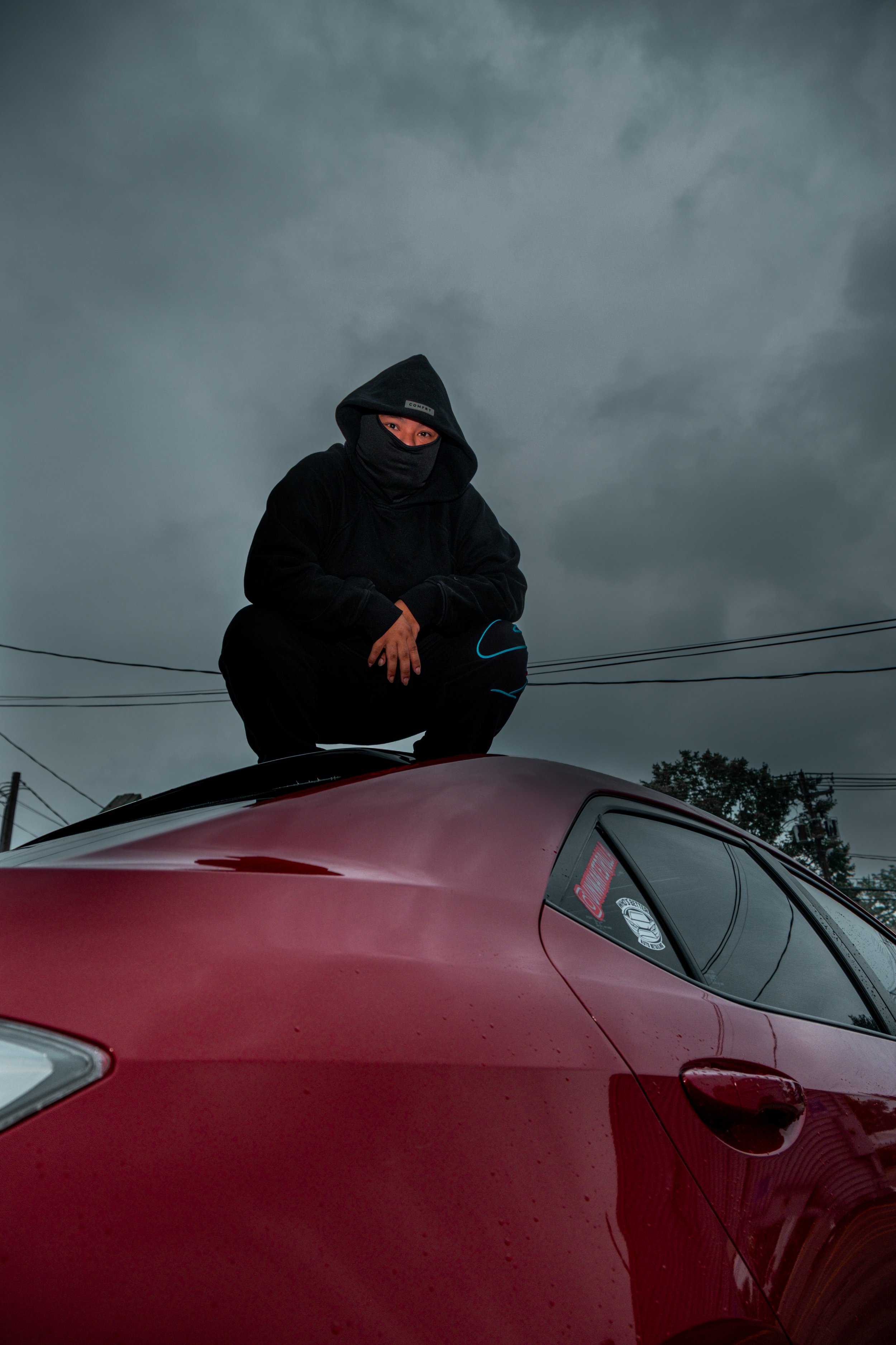 Person wearing a black hooded jacket and face covering squatting on top of a red car under a cloudy sky.