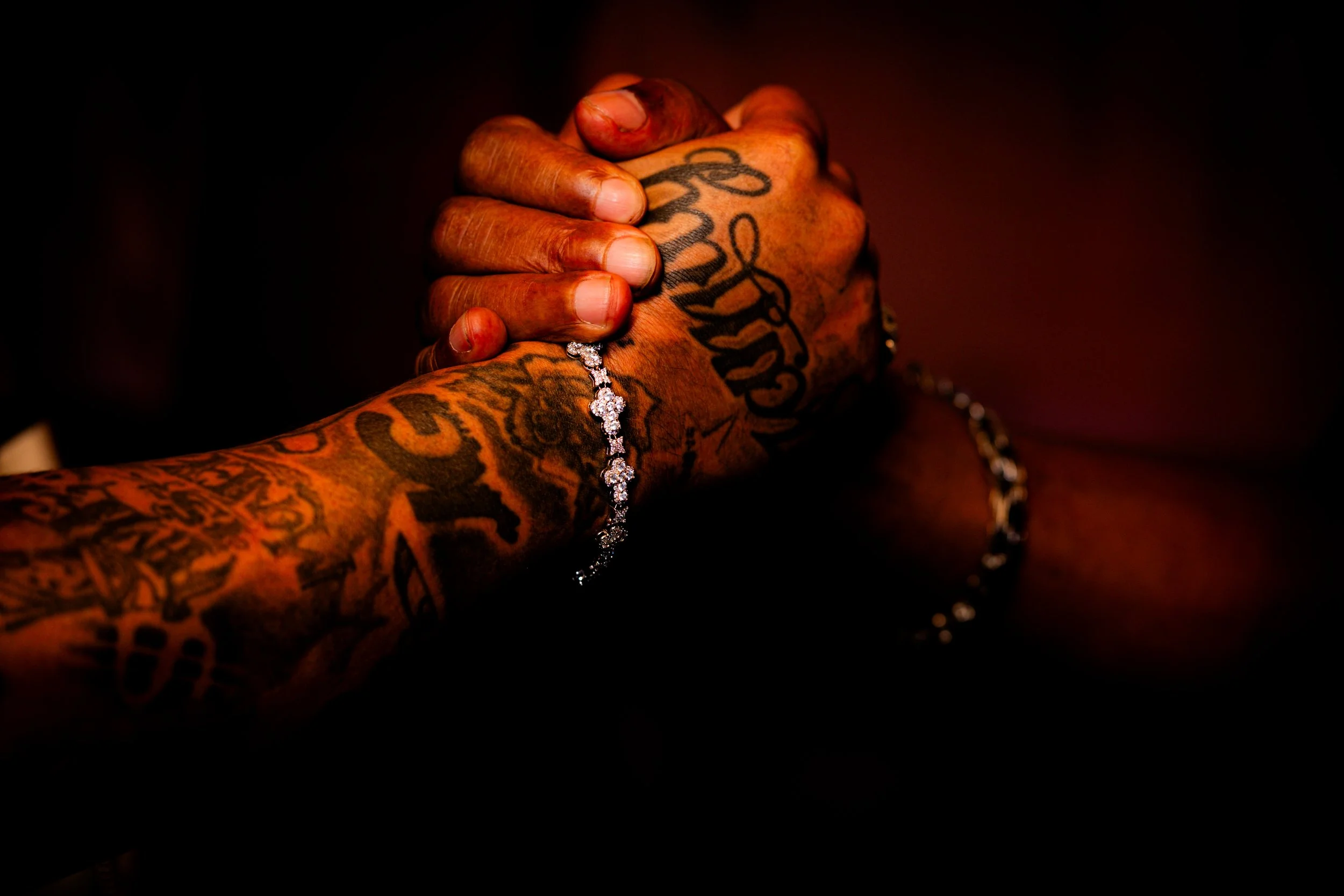 Close-up of a person's tattooed hands clasped together, wearing a diamond bracelet on the wrist, with a dark background.
