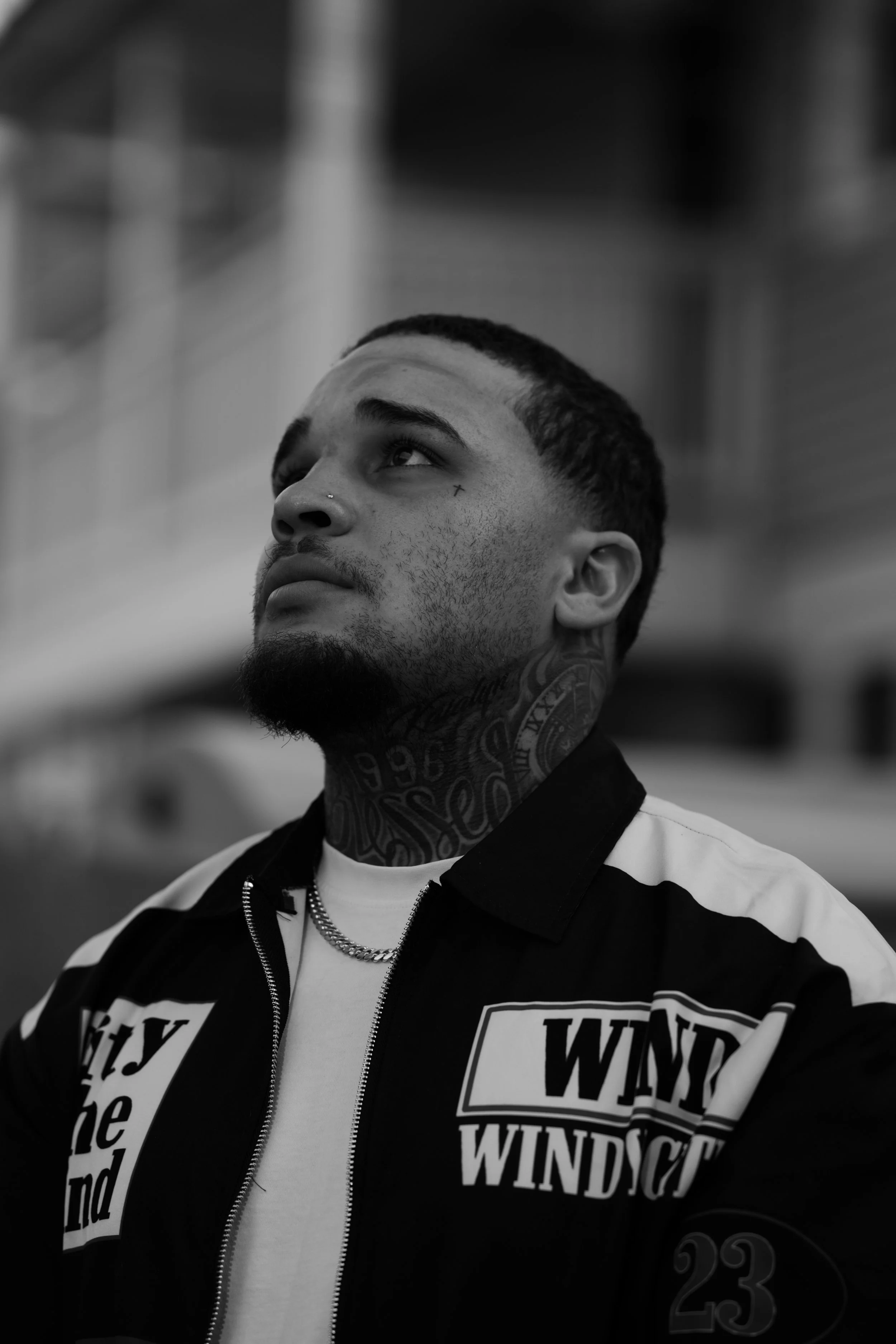 A young man with tattoos on his neck and face, wearing a zip-up jacket with patches and lettering, looking upward against a blurred background.