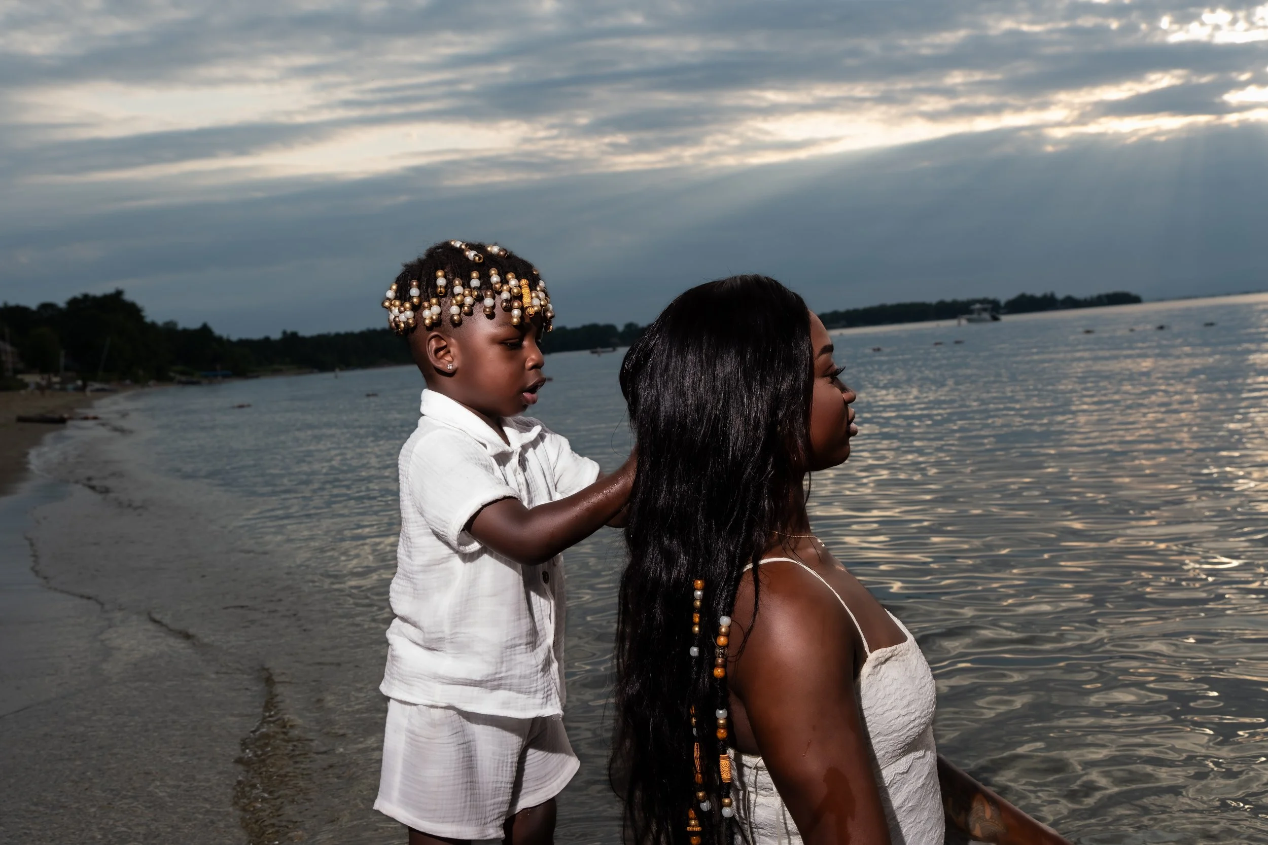 A woman with long black hair and tan skin, sitting by the water, has a young child with tan skin and short hair dressed in white standing behind her, reaching over her shoulder. The child has hair styled with beads and gold accessories. The scene is 