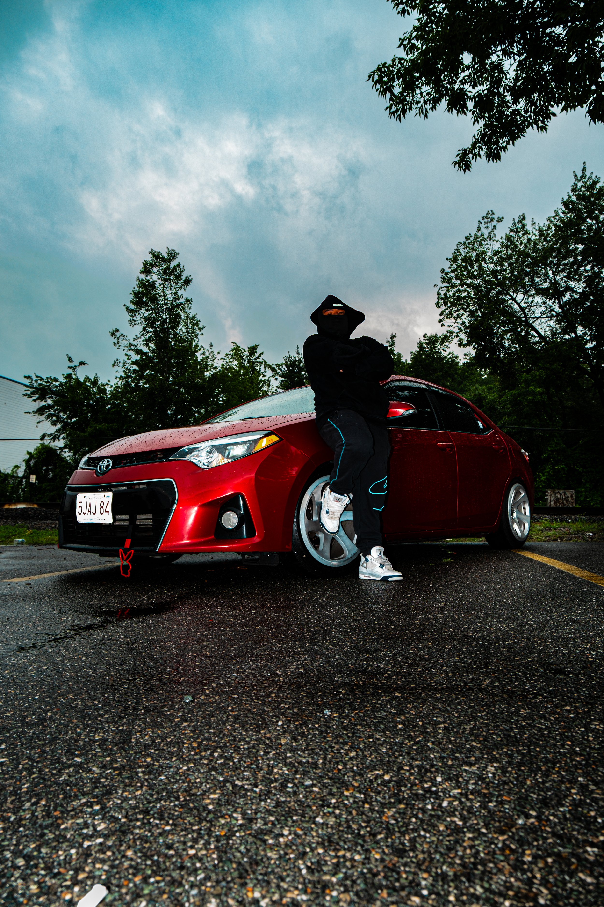 A person dressed in black with a hood and mask standing with crossed arms next to a red Toyota Prius in a parking lot, with green trees and cloudy sky in the background.