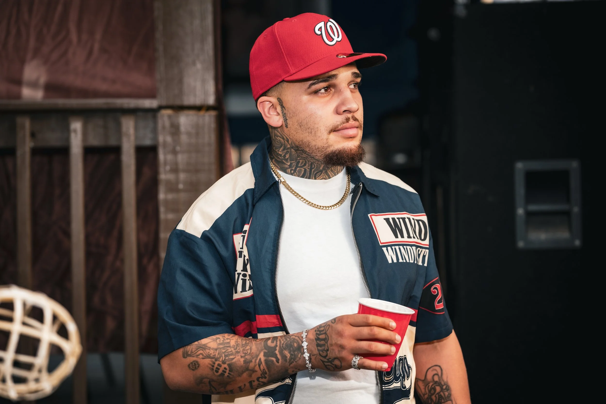 Young man with tattoos on his arms and neck, wearing a red baseball cap with a 'W' logo, a navy blue jacket with patches, a white shirt with graphics, and chains around his neck, holding a red cup, standing indoors near wooden panels and black equipm