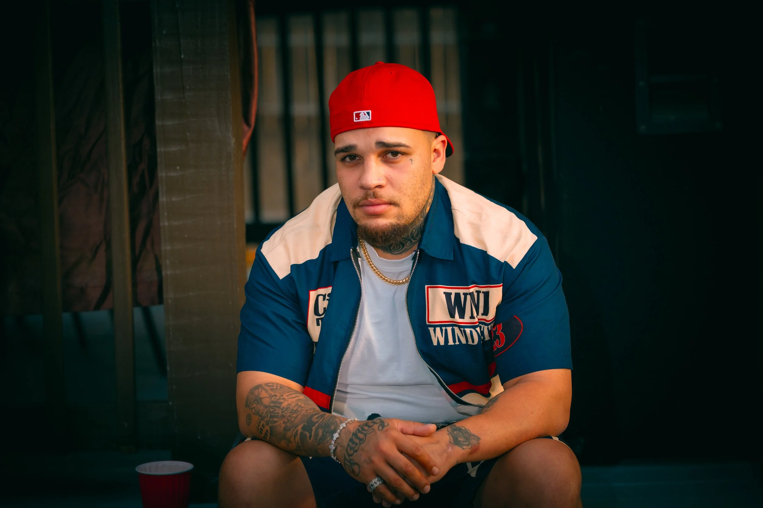 A young man with tattoos on his arm and neck, wearing a red baseball cap backward and a blue jacket with patches, sitting with his hands clasped, looking at the camera in a dimly lit indoor setting.