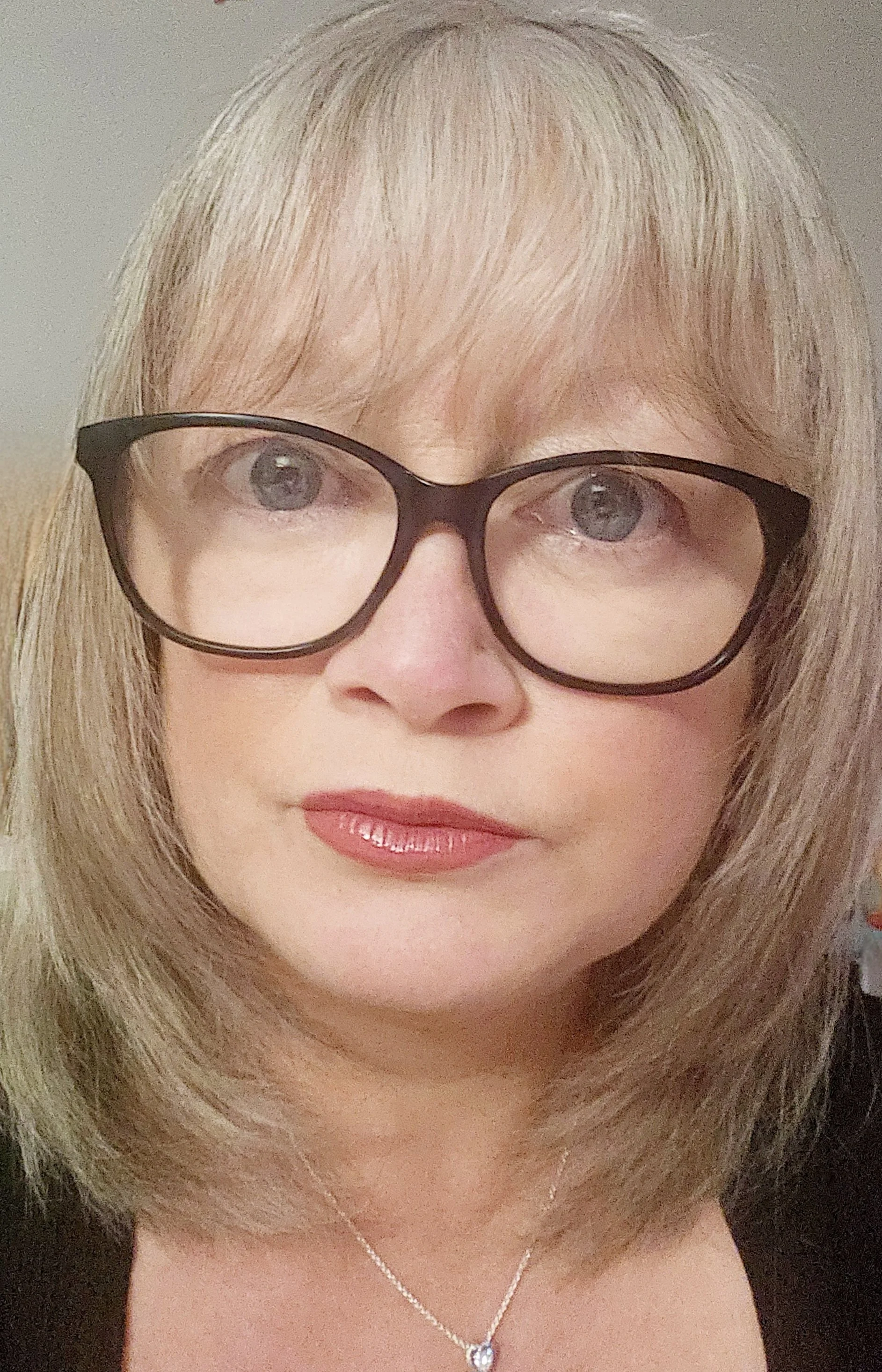 A woman with blonde hair wearing glasses and a silver necklace with a heart-shaped pendant.