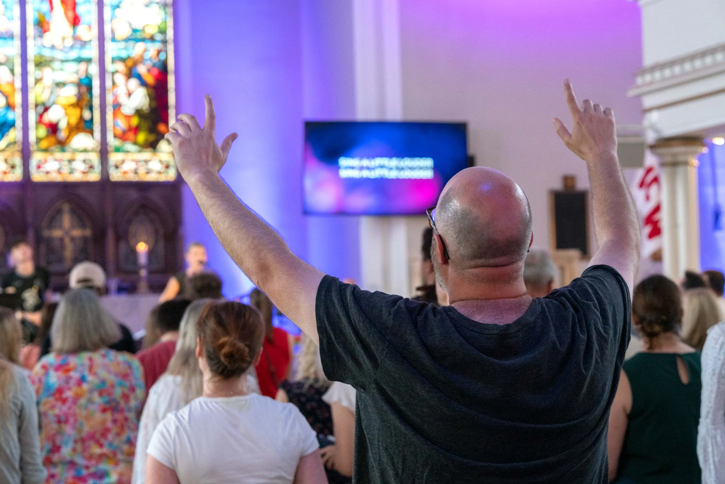 A man with a bald head and glasses raises his hand in a church or gathering with stained glass windows and a large crowd.