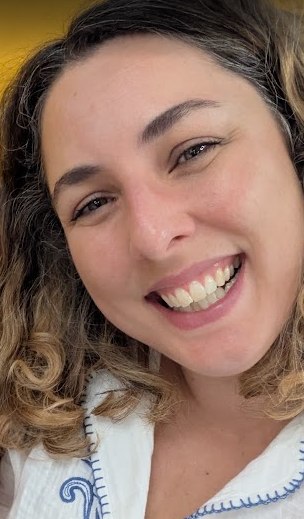 A young woman with curly hair and light skin smiling, showing her teeth, with a light-colored top with blue stitching.