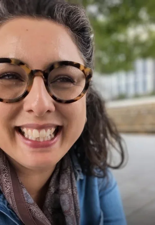 A woman, Tash Hallett, with dark curly hair wearing round tortoiseshell glasses and a patterned scarf, smiling softly outdoors with trees and a stone wall blurred behind her.