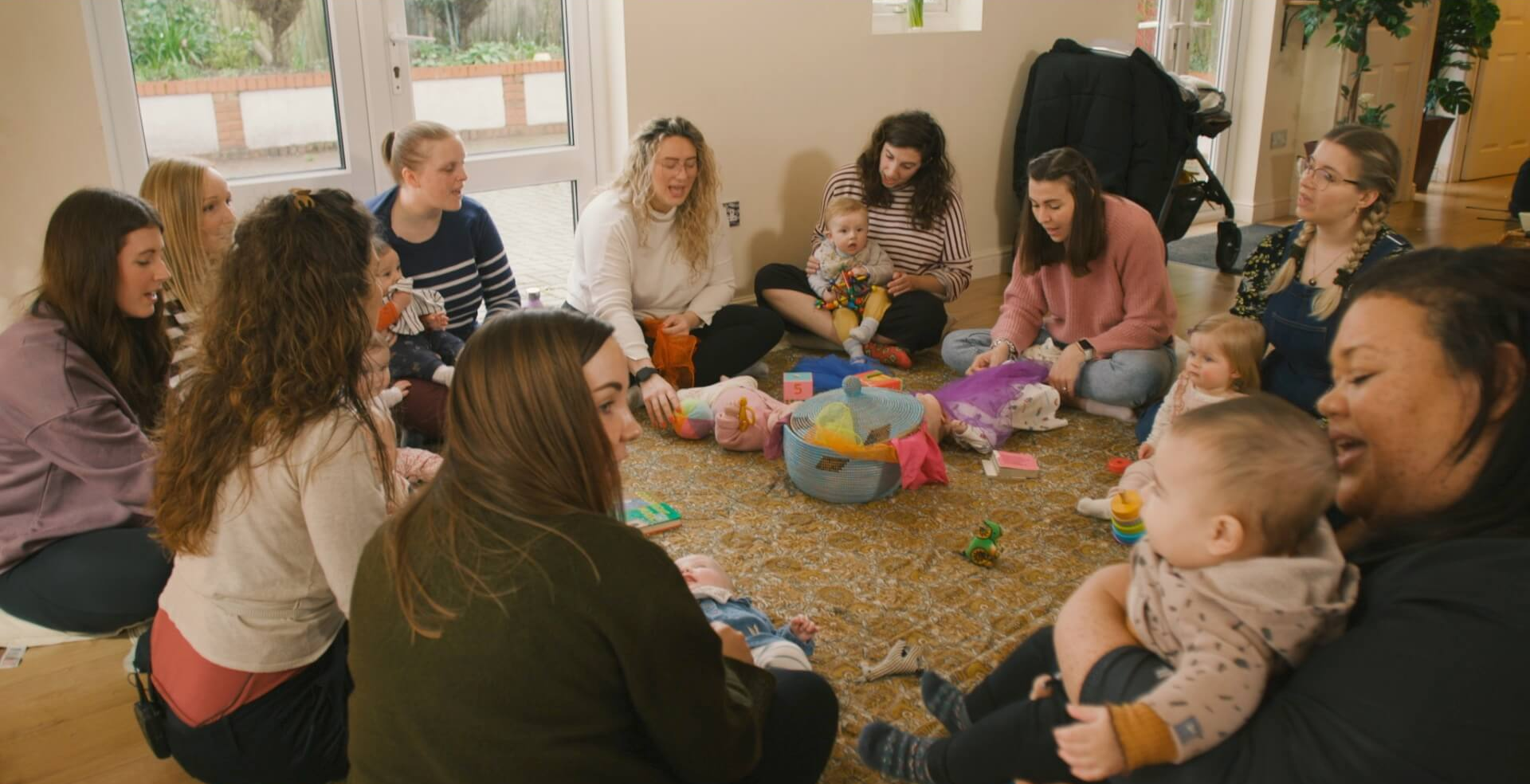 A group of women sitting in a circle on a patterned rug with their babies, singing and playing together in a bright, welcoming room filled with natural light.