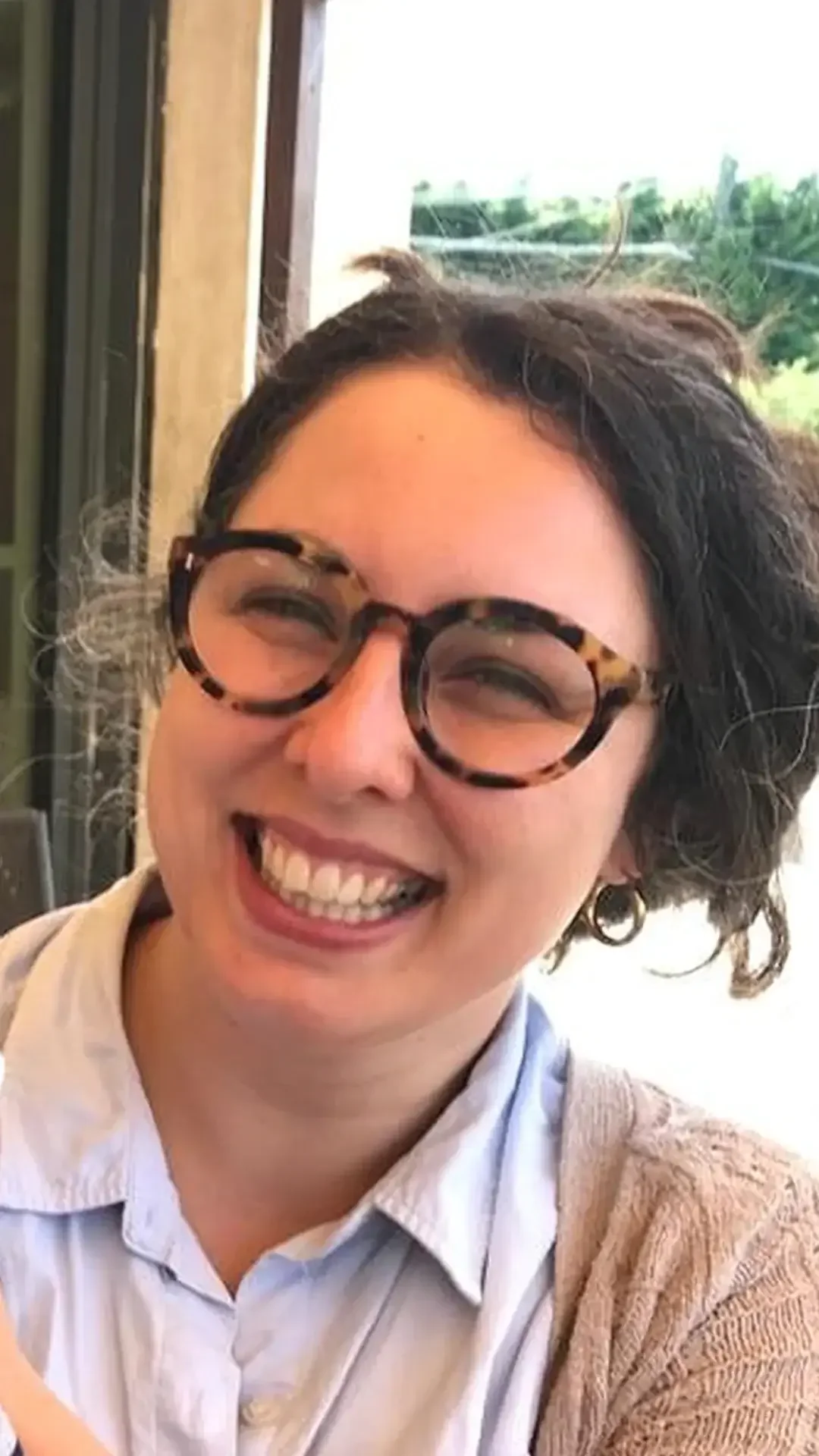 A portrait of Tash Hallett, smiling broadly and laughing with her head tilted slightly. She wears her signature dark curly hair, tortoiseshell glasses, and a light blue shirt layered with a beige cardigan.
