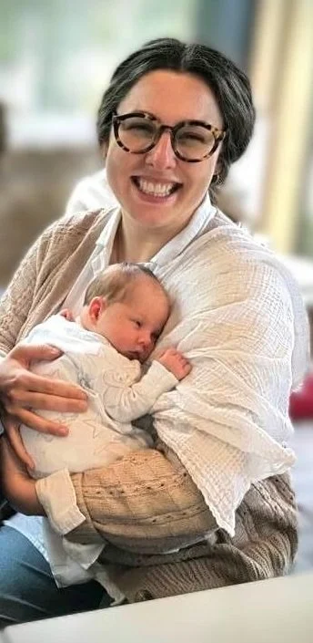 Tash Hallett holding a newborn baby indoors in a bright, relaxed setting.