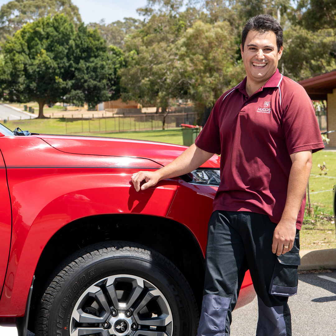 A man in a maroon uniform standing next to a red car in a park, smiling.