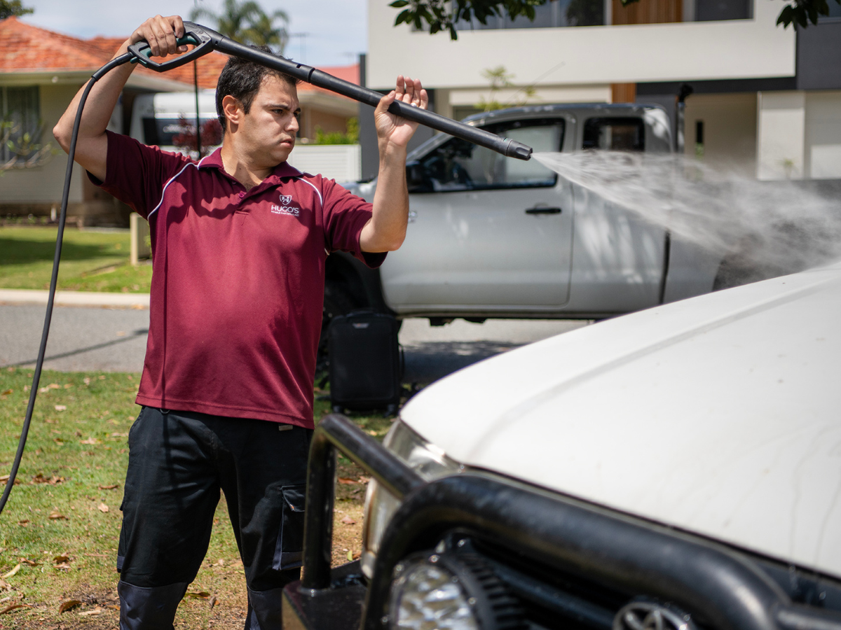 A man in a maroon polo shirt cleaning a white vehicle with a pressure washer outdoors on a sunny day.