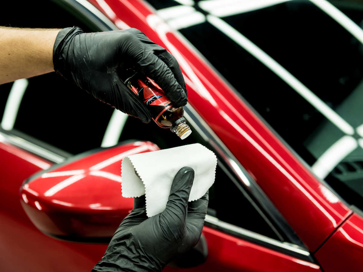 Person wearing black gloves applying a liquid cleaner to a red car's side mirror with a white cloth.