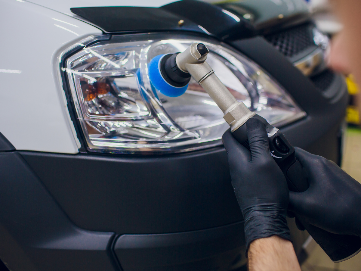 A technician in black gloves is polishing a car's headlight with a power buffer. The car has a white and black exterior.