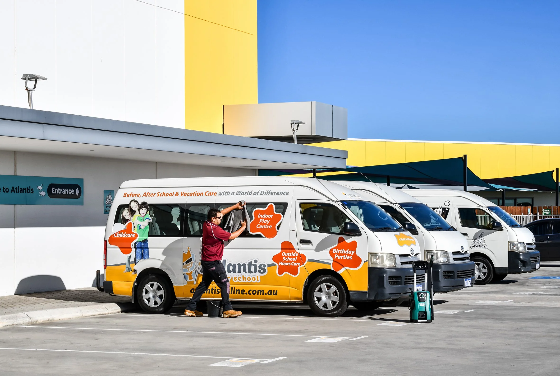 A parking lot in front of Atlantis Preschool, with a row of white vans parked. A man in a maroon shirt is cleaning or organizing one of the vans. The vans have colorful advertisements for preschool services like childcare, birthday parties, and outside school hours care. In the background, there is a modern building with white, yellow, and blue architectural elements, and a clear blue sky.