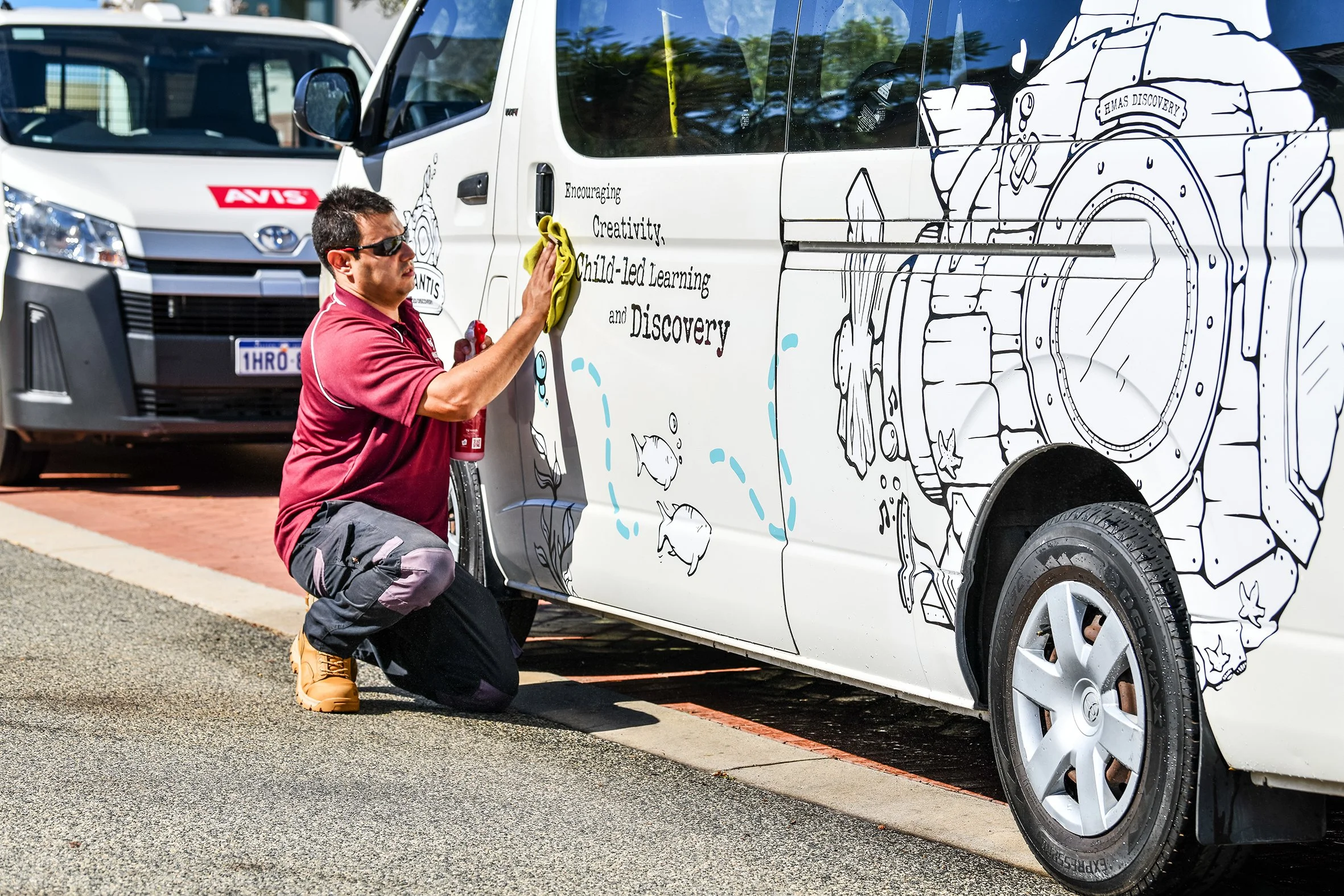 A person kneeling on the ground, cleaning the side of a white van with black illustrations and text, while wearing a red shirt, black pants, and tan boots.