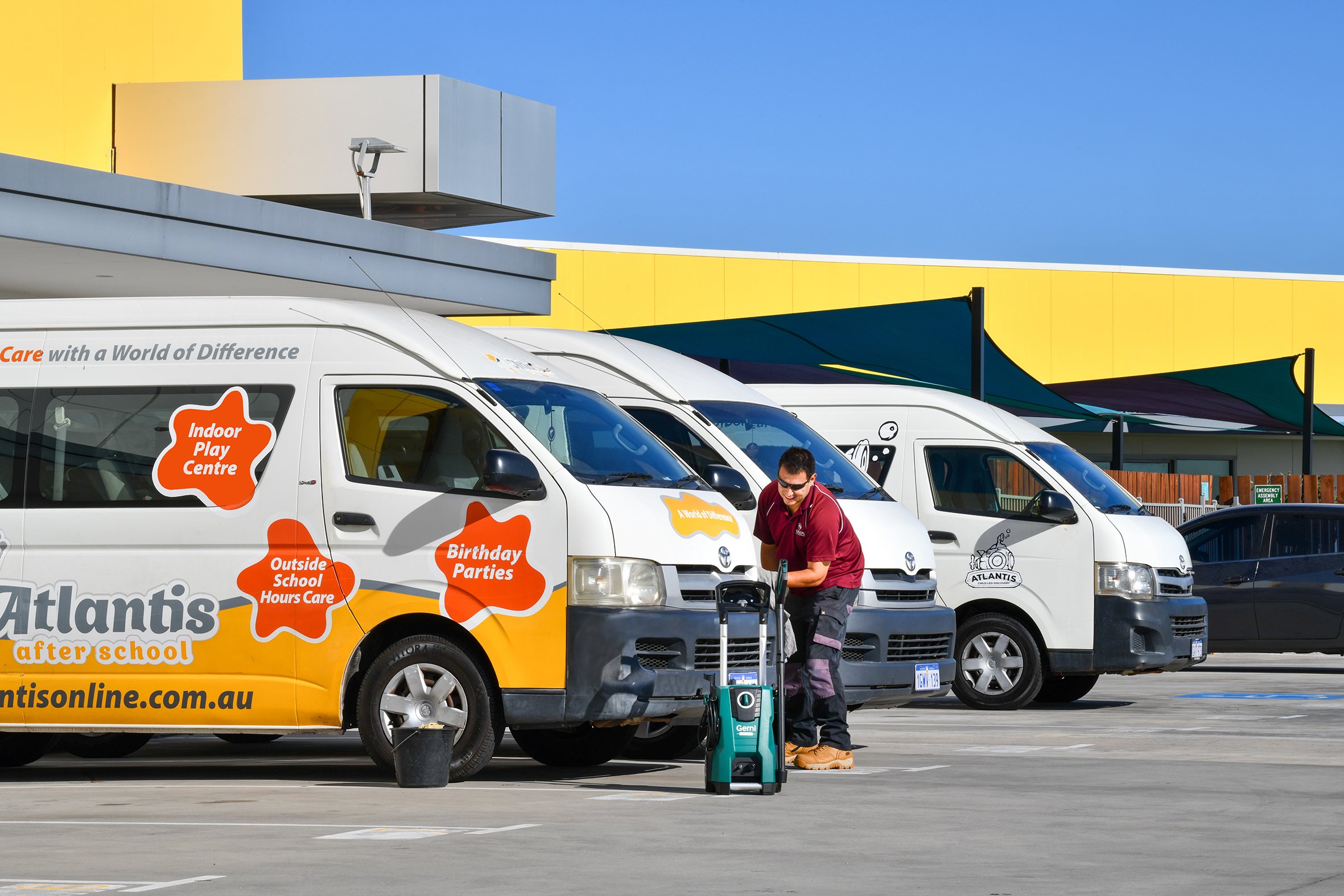 A man standing next to a row of white vans with colorful graphics and text on the side, parked in an outdoor lot under a yellow and blue building. The man appears to be working or organizing equipment.
