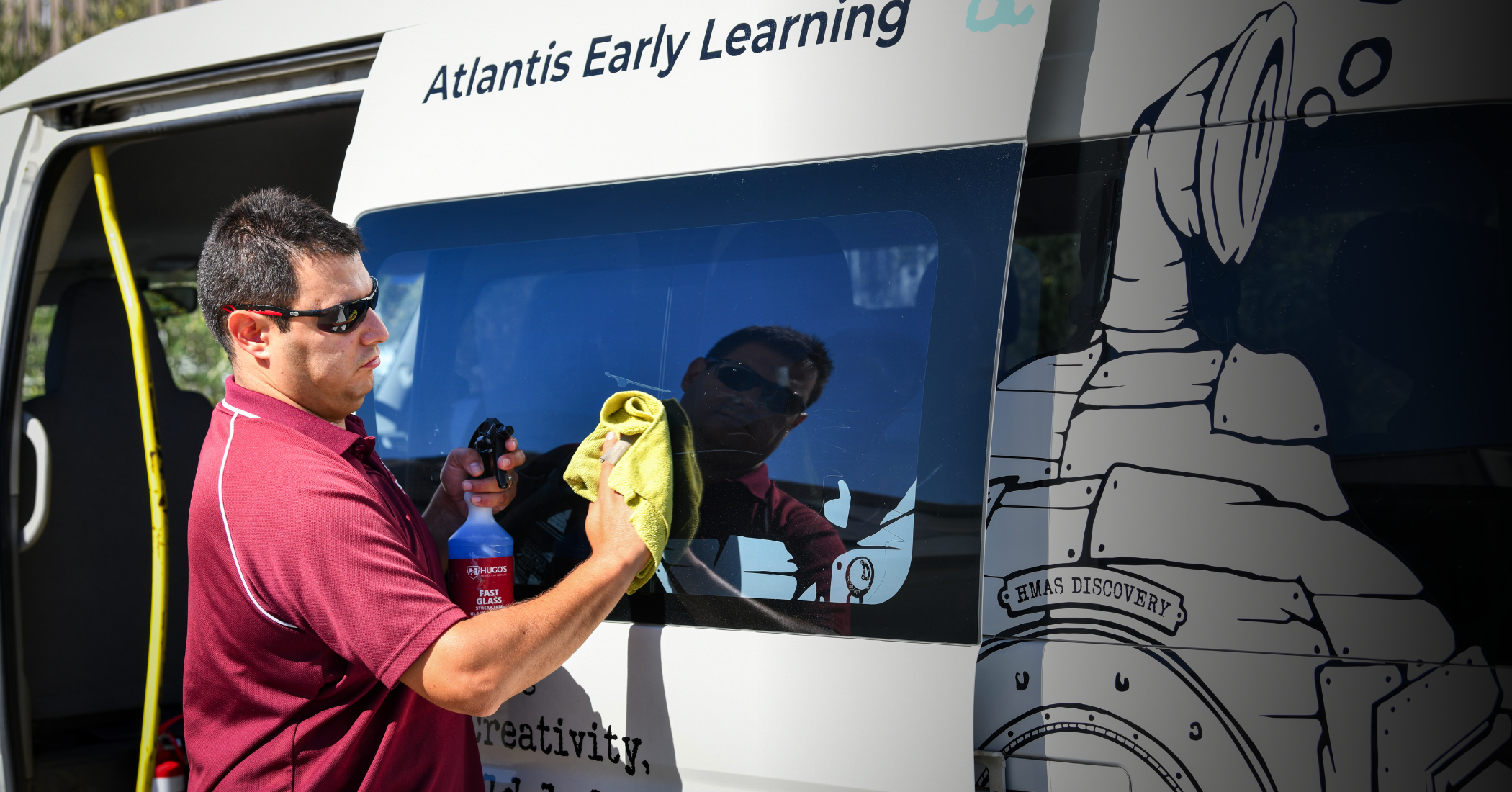 A man cleaning the side window of a vehicle labeled 'Atlantis Early Learning' with a yellow cloth, wearing sunglasses and a maroon polo shirt, holding a spray bottle in his left hand, with a reflection of himself in the window.