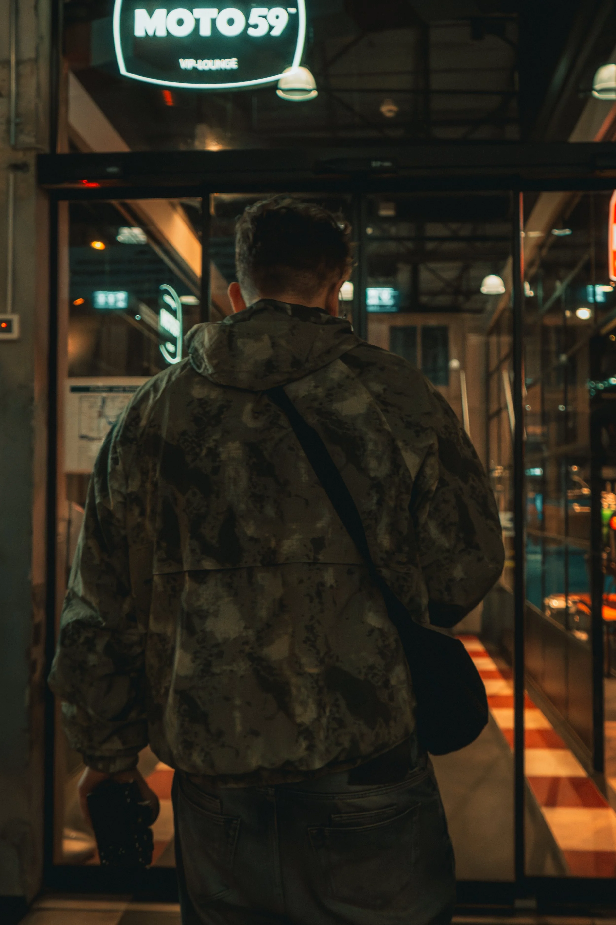 Ein Mann aus hinterer Perspektive in einer Jacke an einem Fahrstuhl. Im Hintergrund ein beleuchtetes Schild mit der Aufschrift 'MOTO 59 VIP-LOUNGE'.