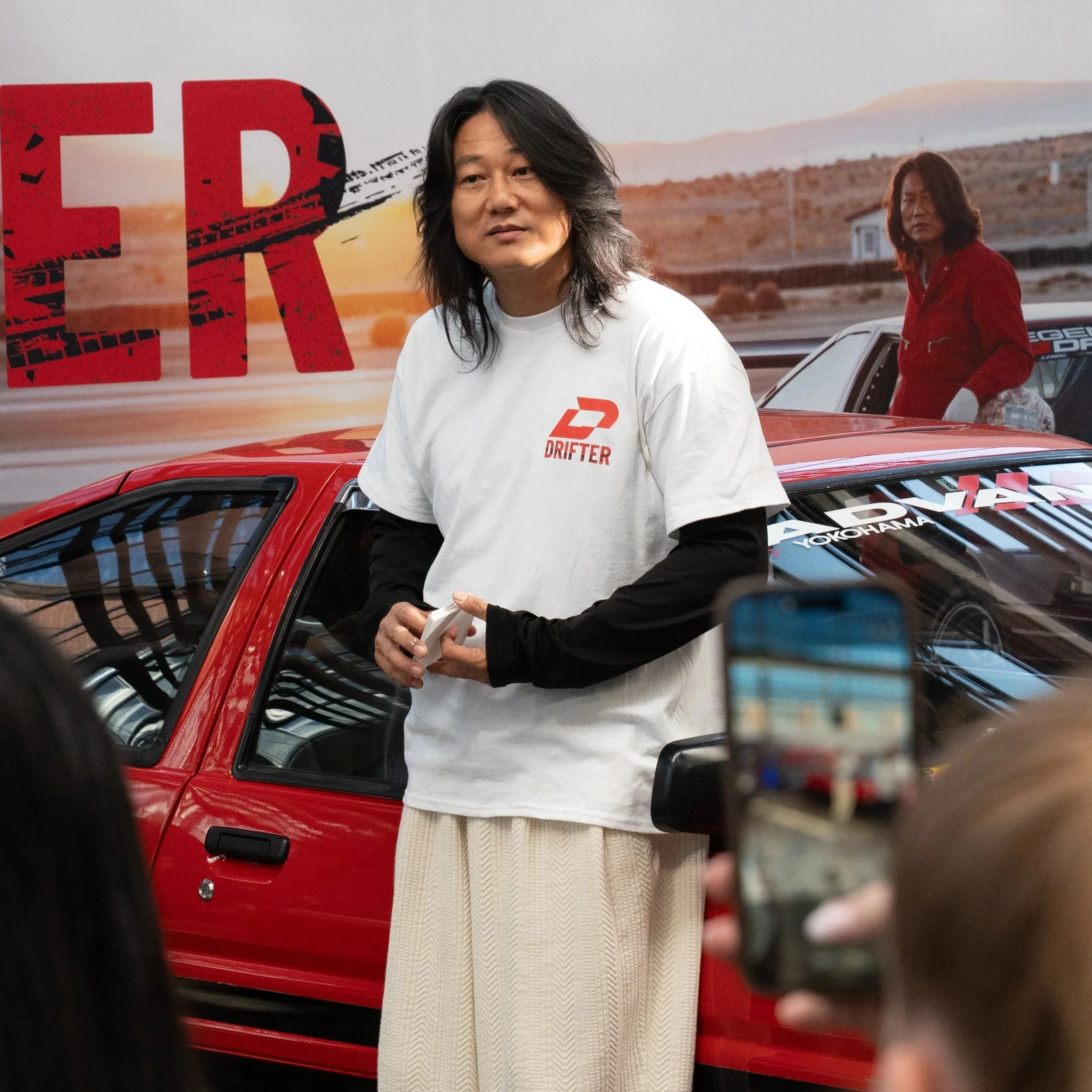 Ein Mann mit langen dunklen Haaren steht vor einem roten Auto, trägt ein weißes T-Shirt mit einem roten Logo, im Hintergrund ist eine ländliche Landschaft bei Sonnenuntergang. Ein zweiter Mensch im roten Jacke steht neben dem Auto.