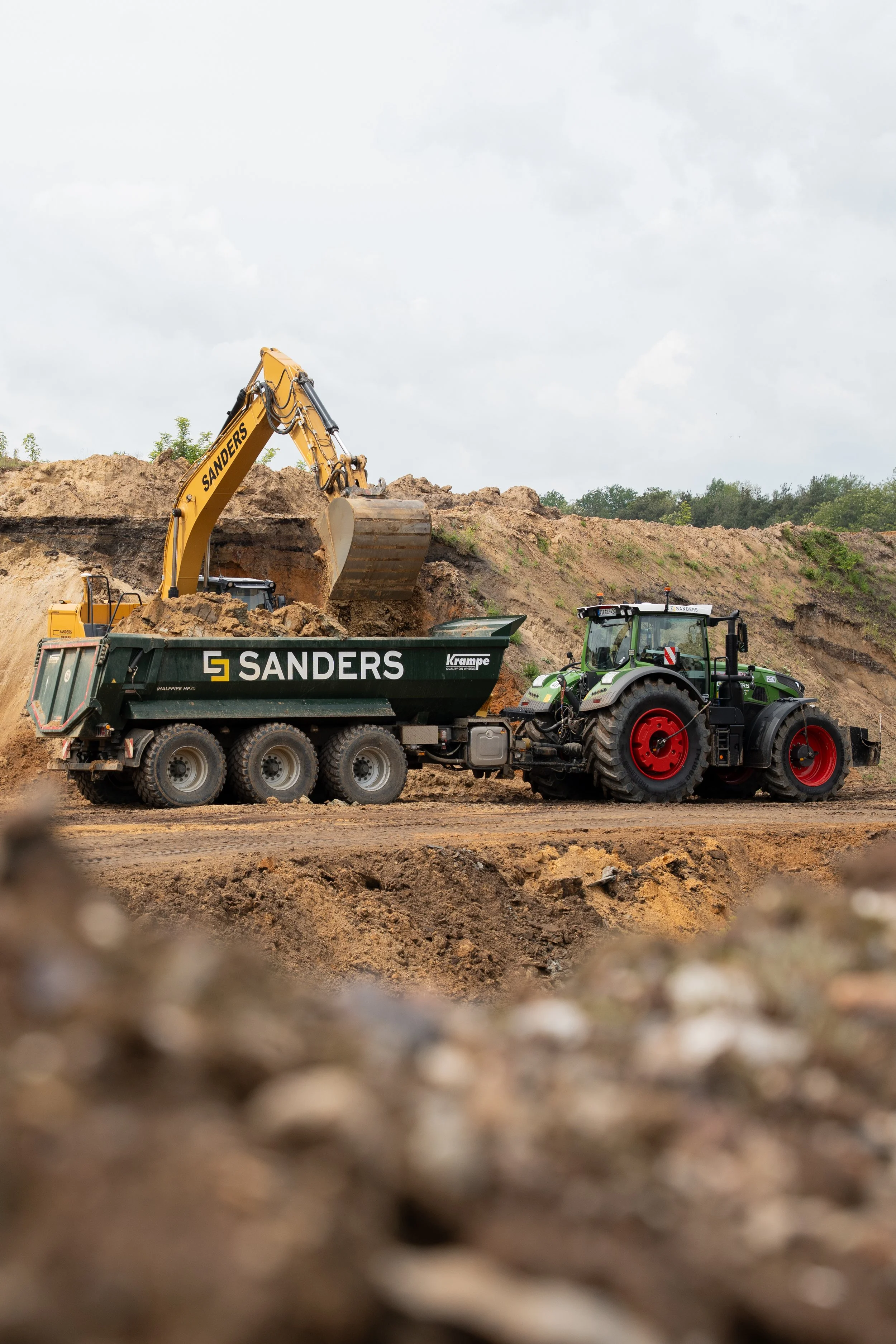 Baustelle mit Bagger und Traktor beim Aushub, Sanders-Marke sichtbar, auf einer Erdbaufläche mit Hügel im Hintergrund.