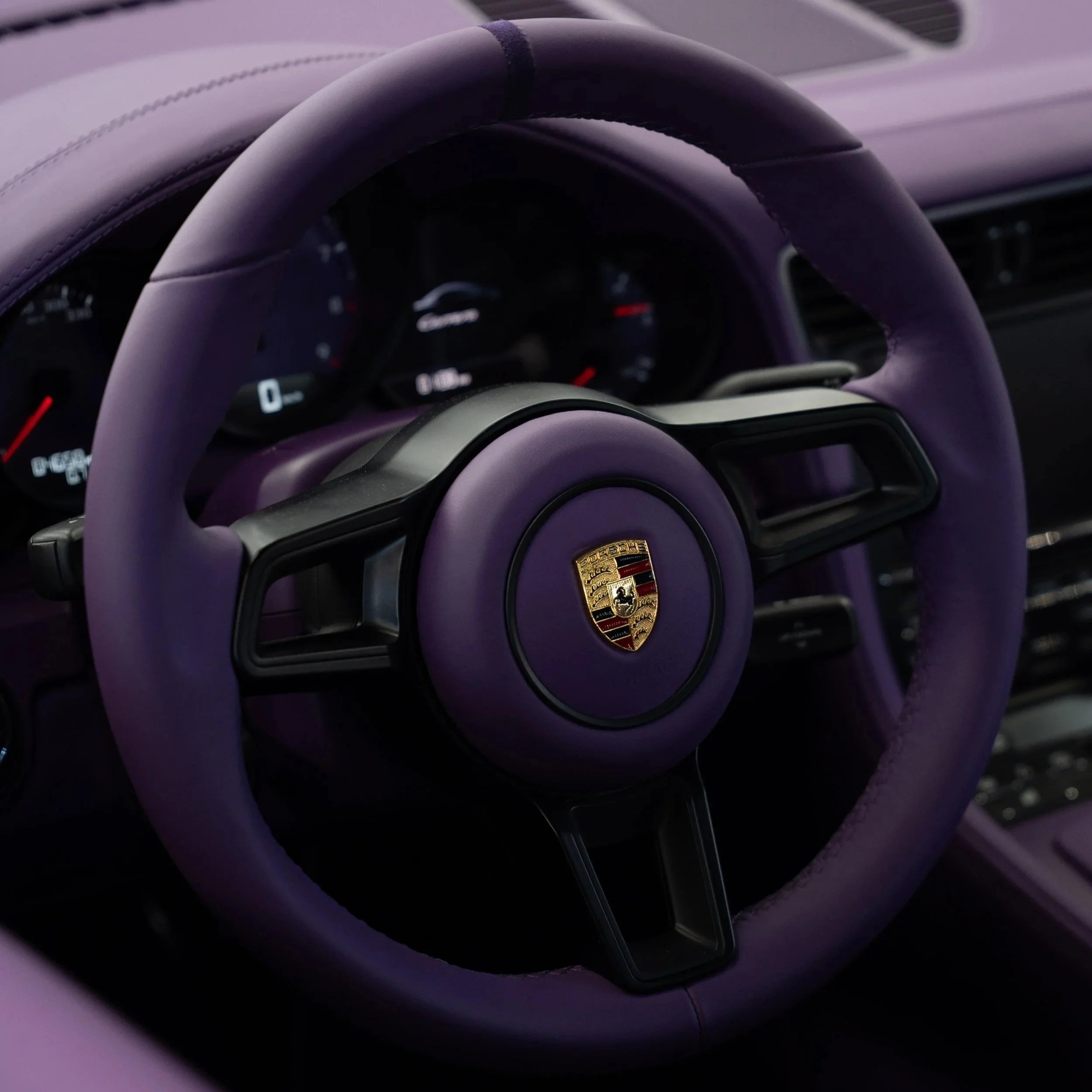 Nahaufnahme eines Lenkrads eines Porsche-Fahrzeugs mit schwarzem Porsche-Logo in der Mitte, umgeben von einem lila Lederbezug.