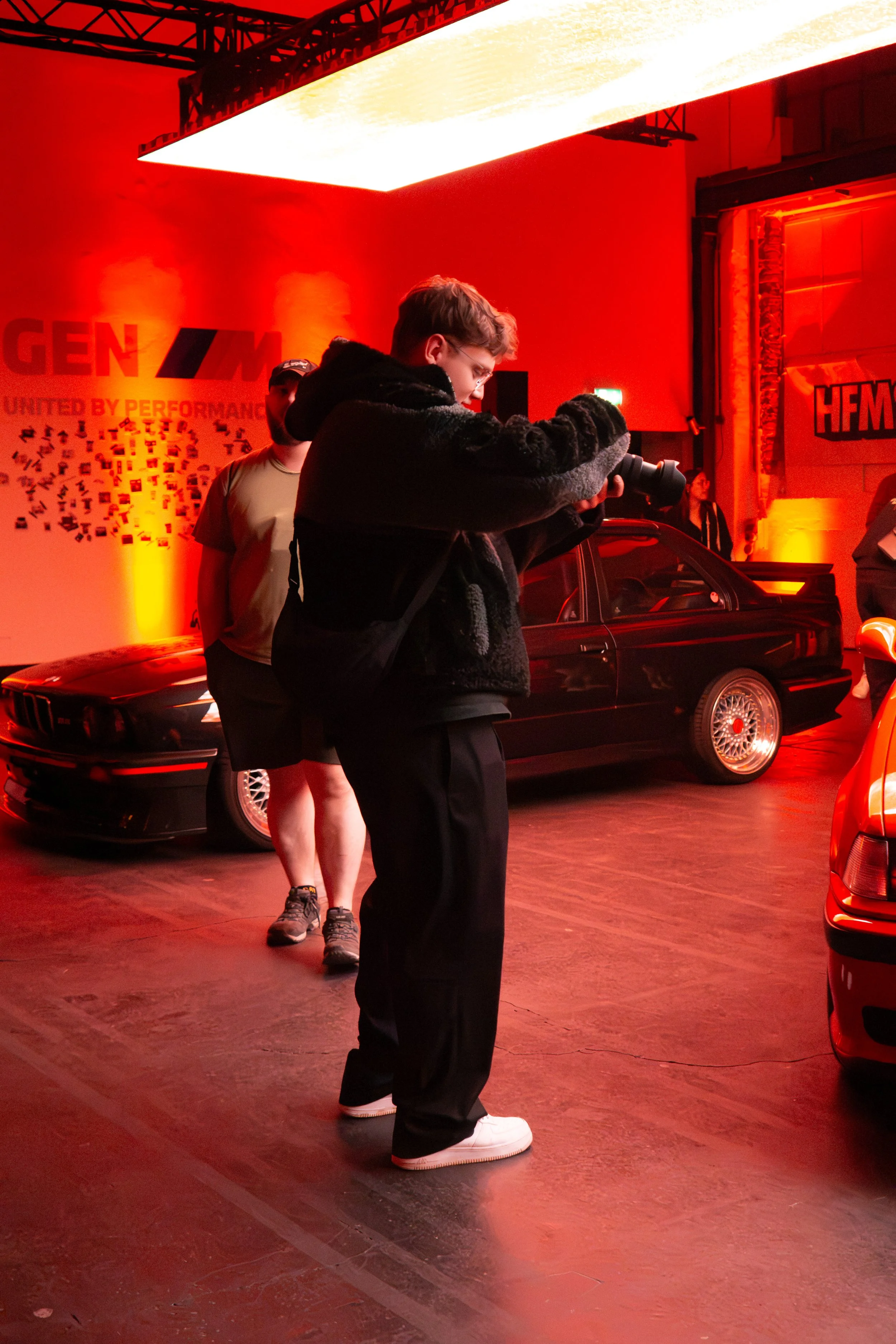 Junge Mann fotografiert in einer Indoor-Auto-Ausstellung, bei warmer roter Beleuchtung, mit klassischen Sportwagen im Hintergrund.