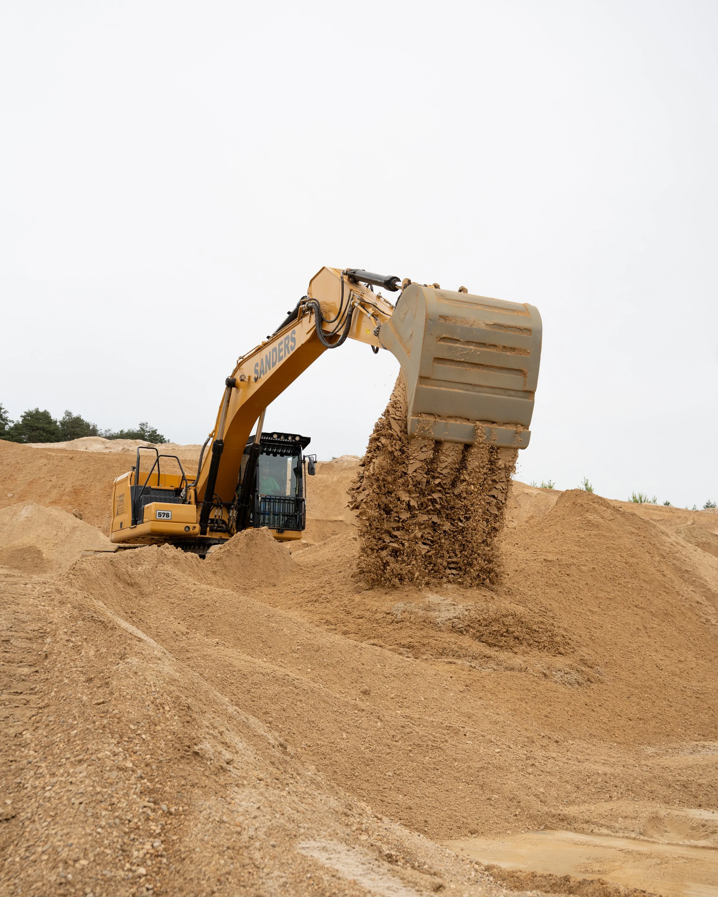 Ein Bagger hebt Erde in einem Baufeld bei einem Bauprojekt. Der Himmel ist grau und bewölkt.