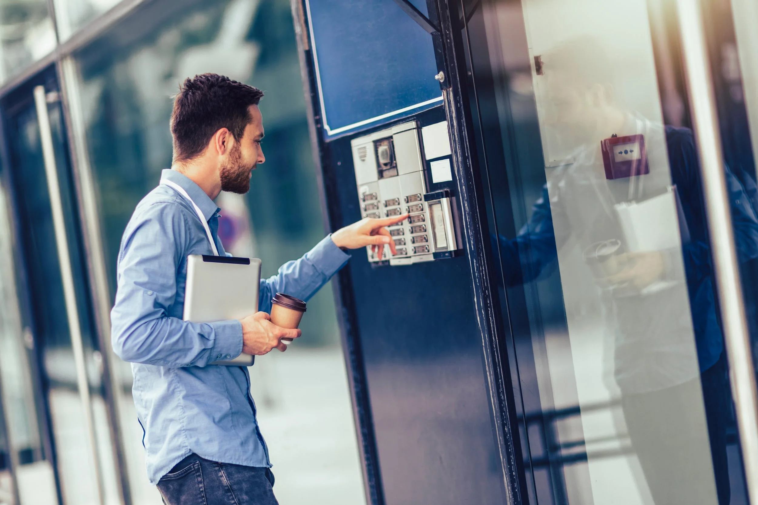 A man with dark hair and a beard wearing a blue shirt using a pay phone outside a building, holding a coffee cup in his left hand and a laptop under his arm.