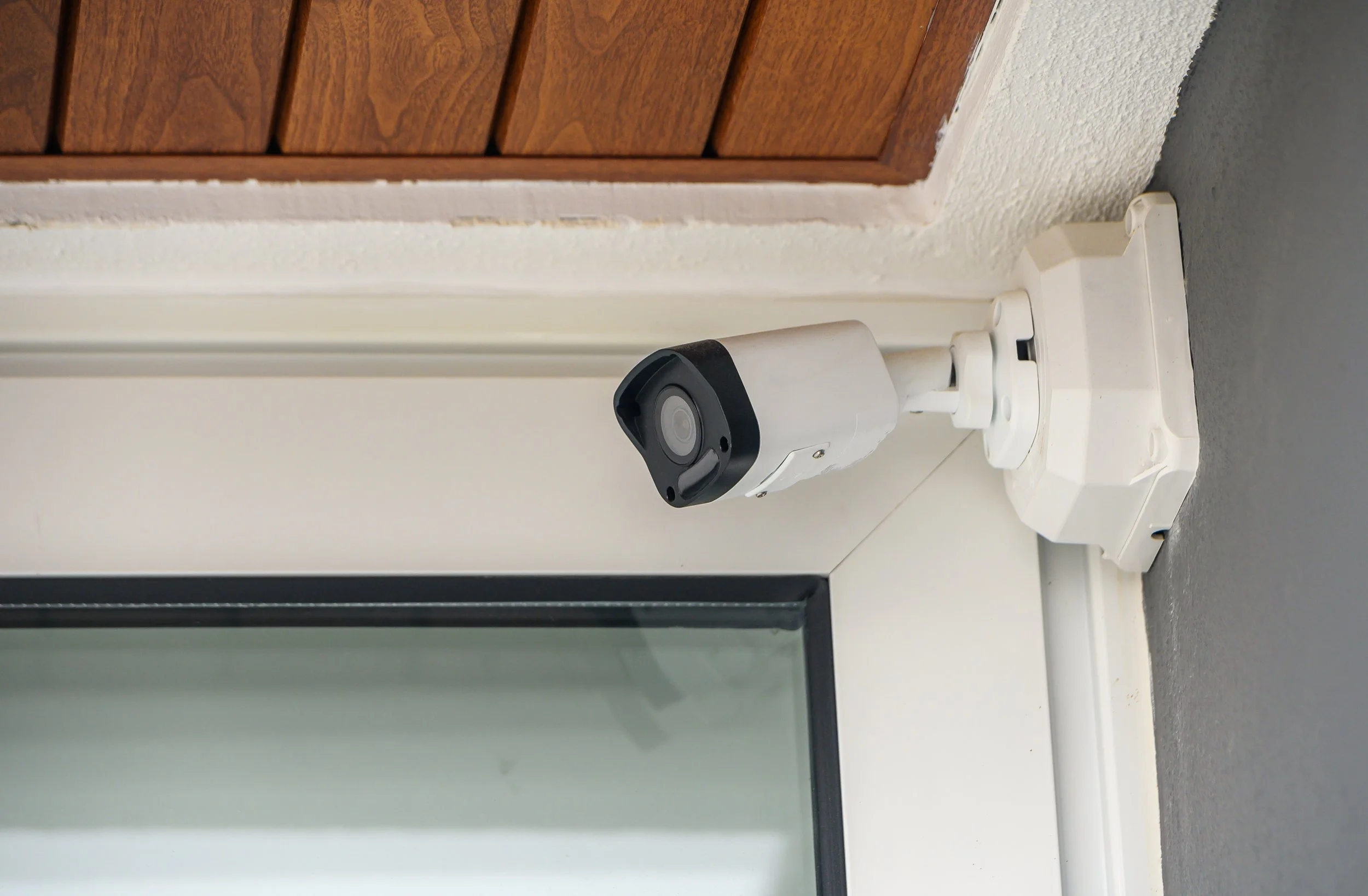 A white security camera mounted on a wall near a door with wooden panels, adjacent to a textured white ceiling and a gray floor.