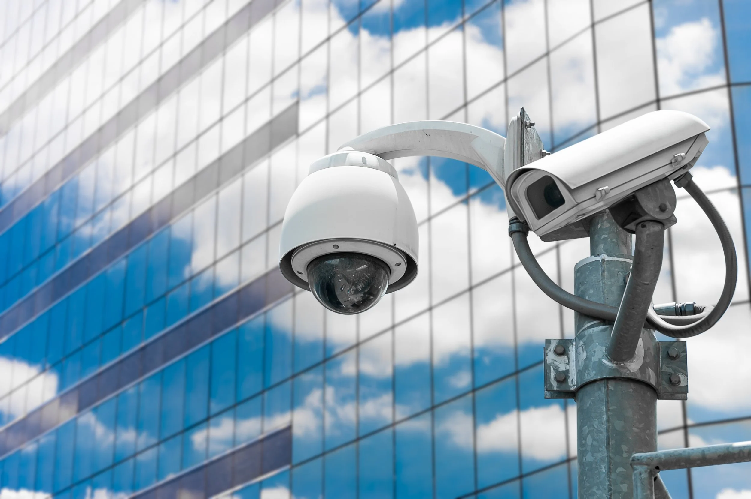 Surveillance camera mounted on pole against sky and reflection of clouds on glass building.