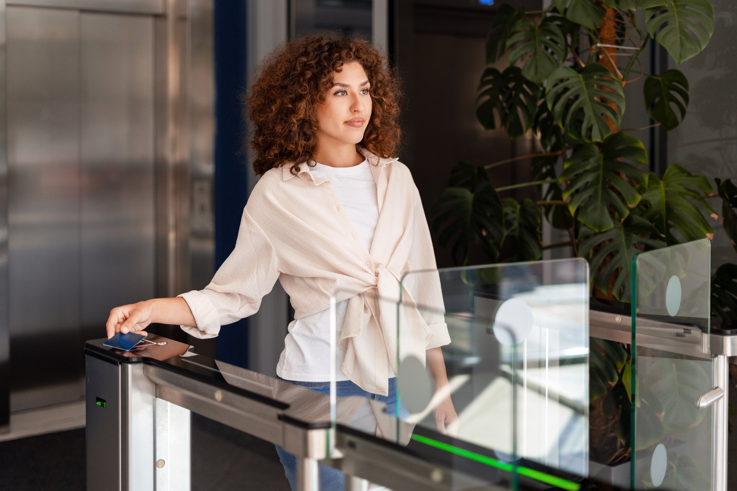 A young woman with curly brown hair standing at a reception desk in a modern building, holding a smartphone, with a large green plant in the background.