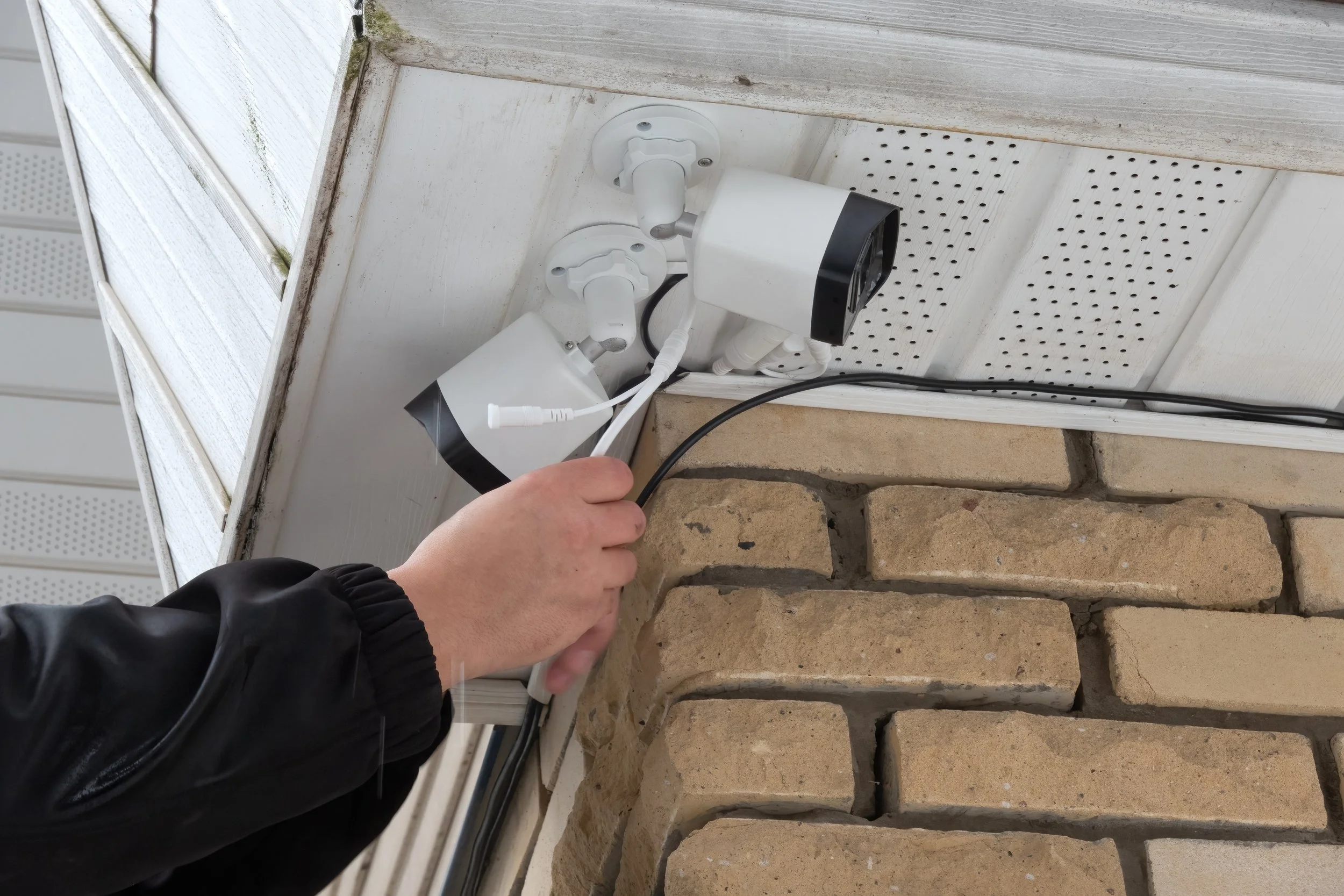 Person's hand connecting outdoor security camera wires near a house with brick and white siding.