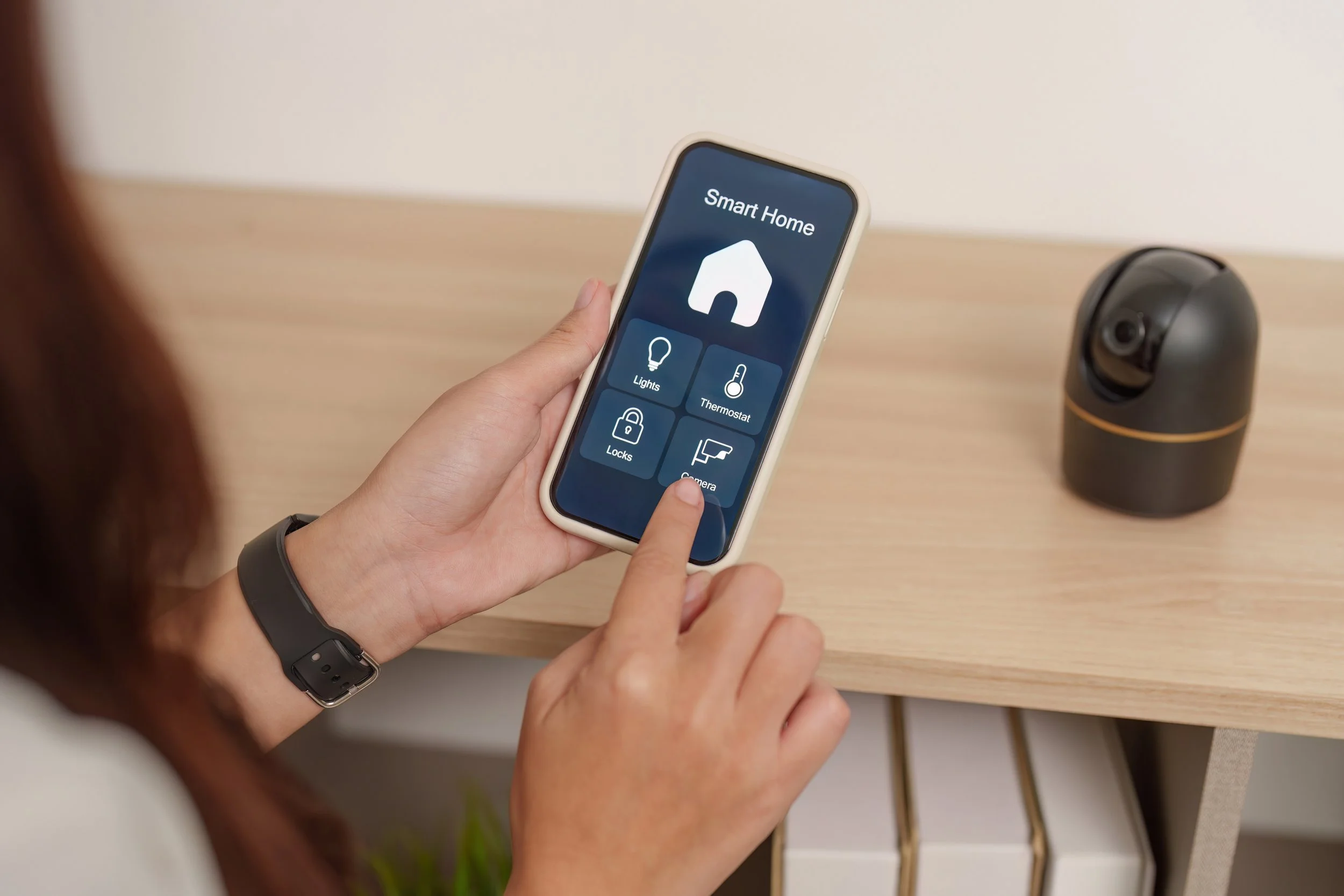 Person holding a smartphone with a smart home app on the screen, showing options for lights, thermostat, locks, and camera, on a wooden surface with a security camera nearby.