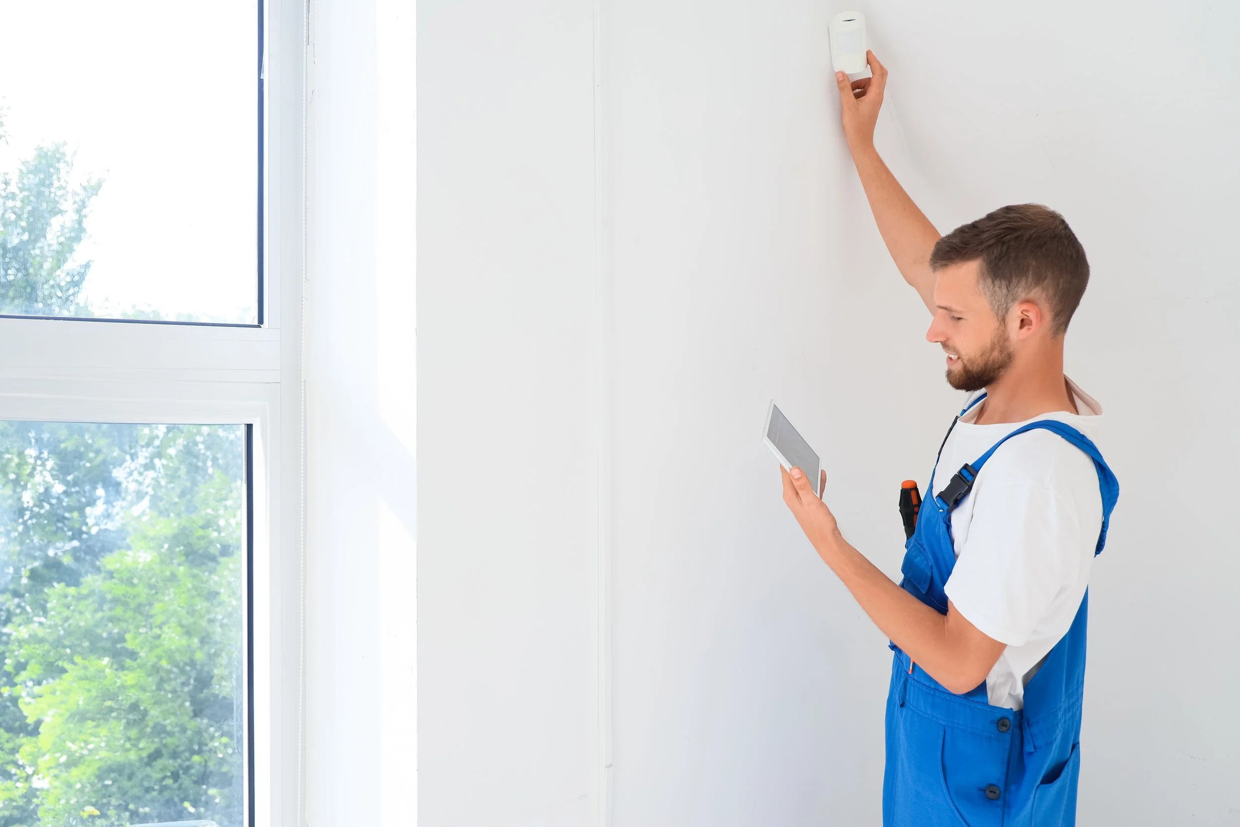 A technician stands inside a bright room with large windows, holding a tablet in one hand and adjusting a motion sensor on the wall with the other hand.