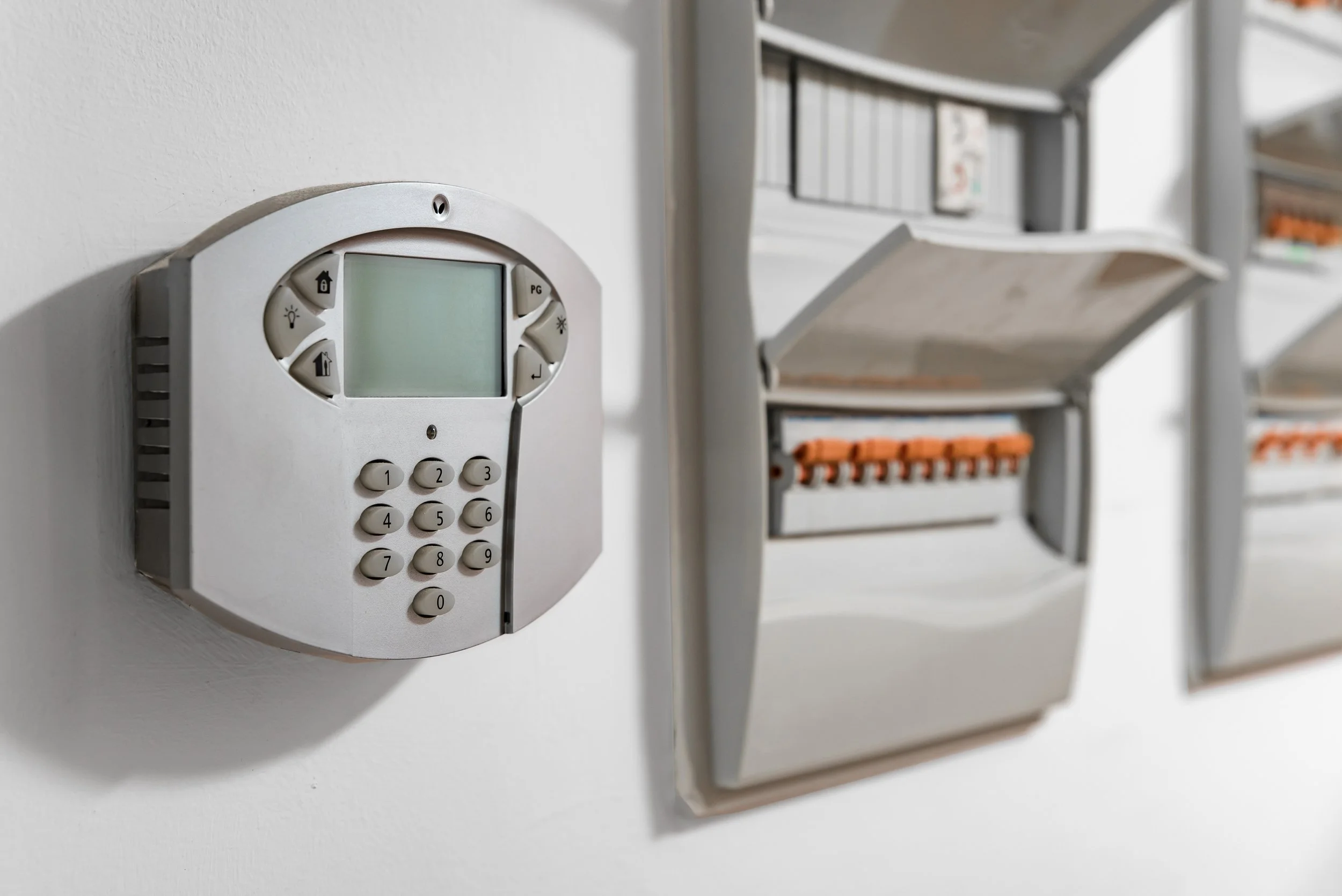 An electronic security system keypad with a display screen mounted on a white wall, with an electrical circuit breaker panel nearby.