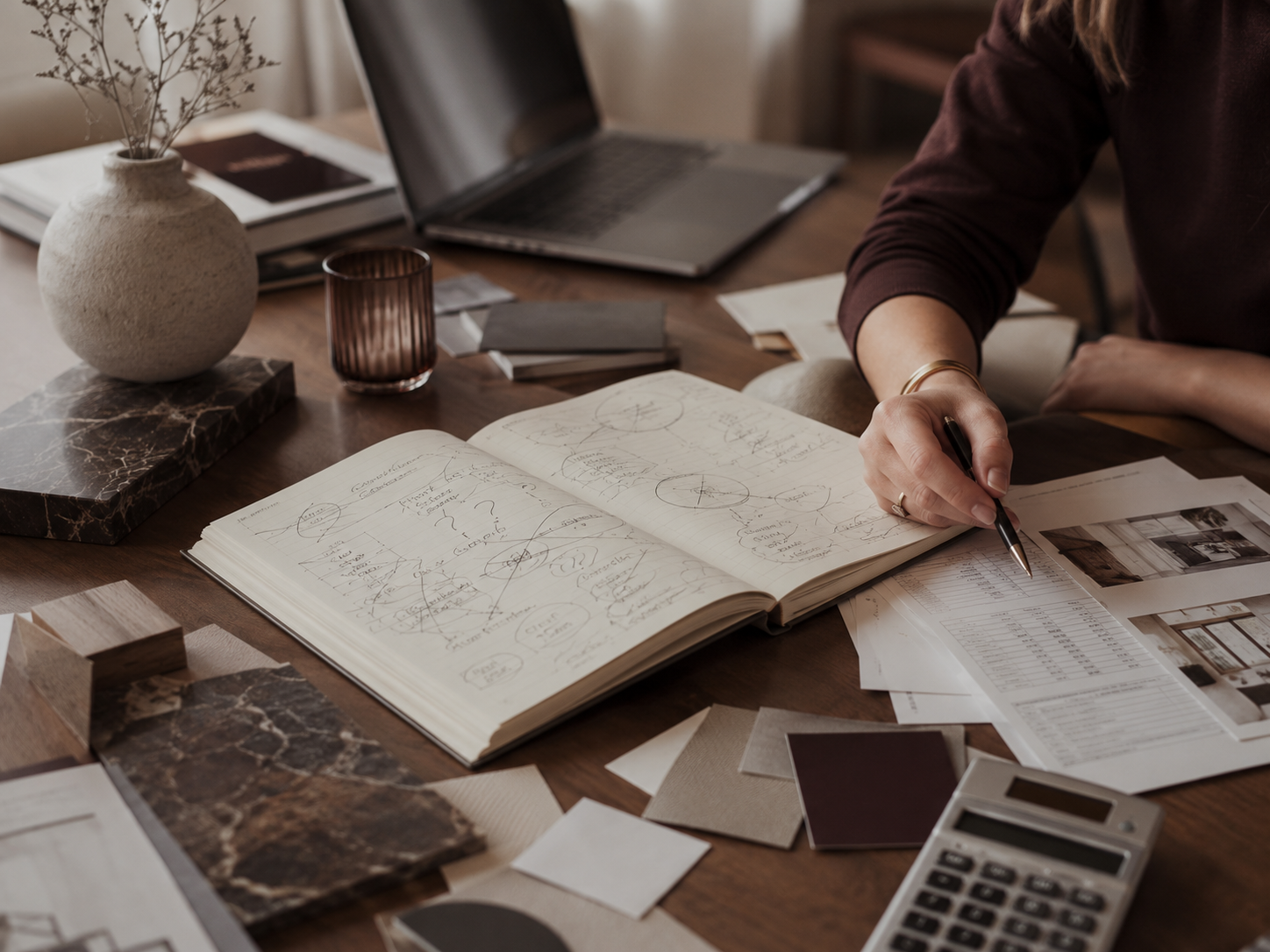 A person working at a wooden desk with an open notebook filled with diagrams and notes, surrounded by documents, a laptop, a vase with dried flowers, and design samples.