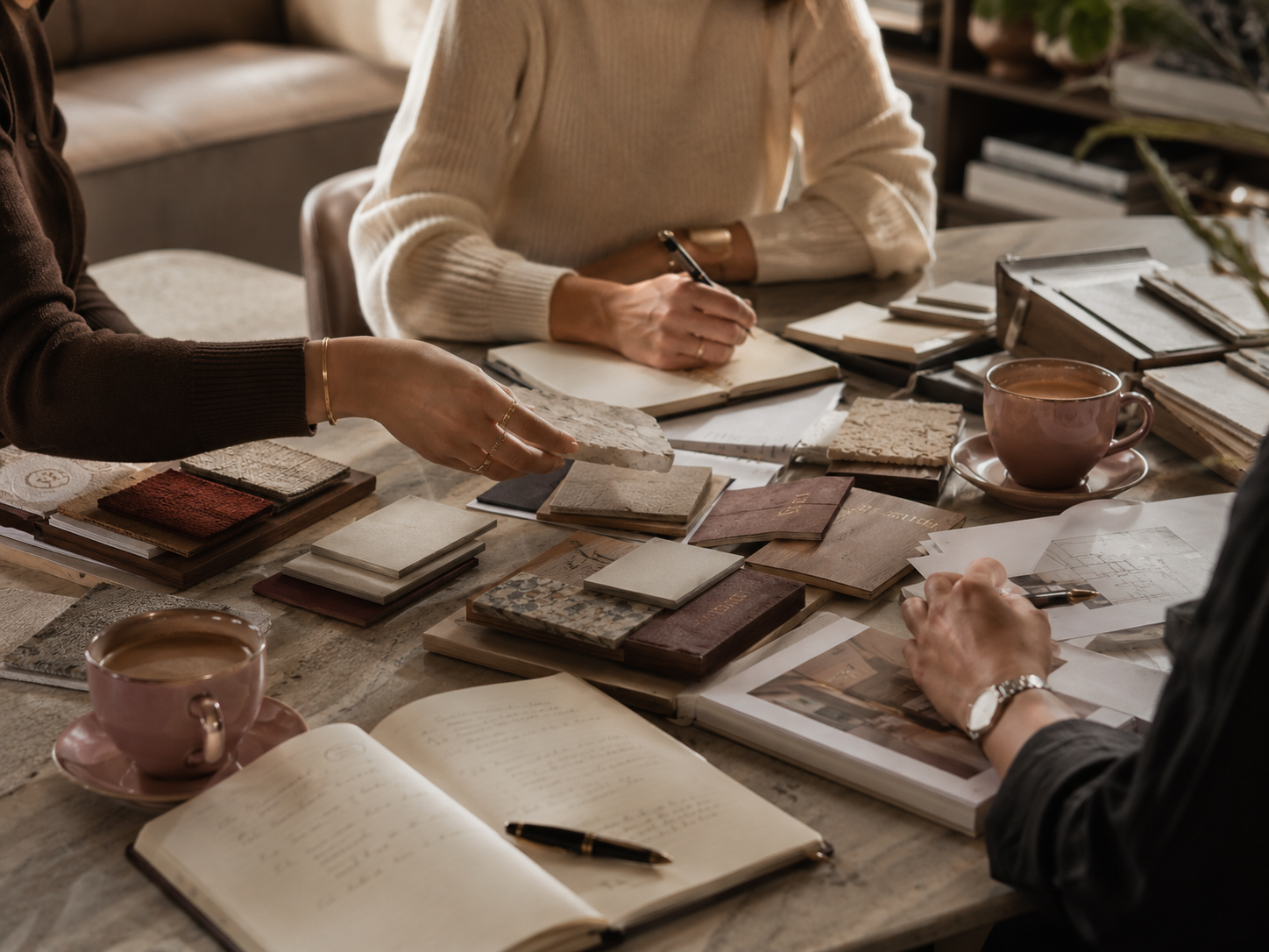 People working on interior design or architecture project with tile samples, books, notebooks, and coffee on a wooden table.