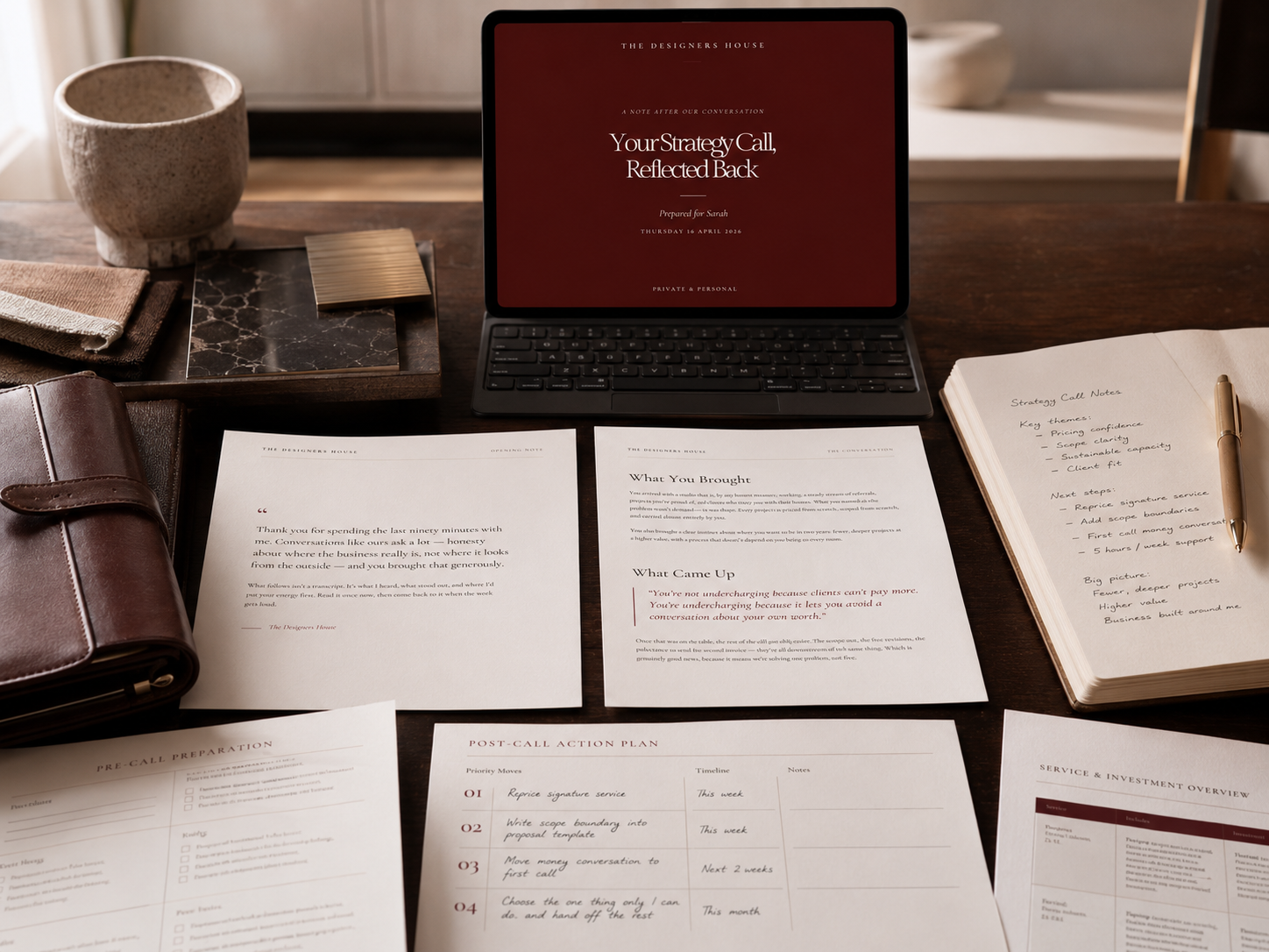 A workspace with a laptop, printed documents, a notebook with handwritten notes, a pen, a leather planner, a tray with fabric samples and a marble tile, and a mug on a wooden table.
