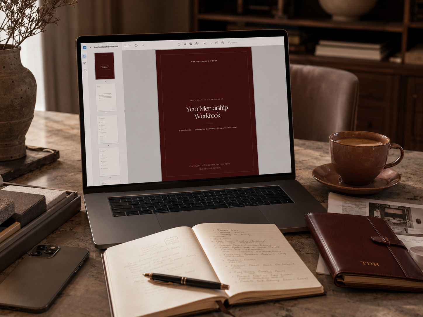 A laptop on a wooden desk displaying a mentorship workbook cover page. The desk has a brown notebook with initials 'TDH', an open journal, a pen, a smartphone, a cup of coffee on a saucer, and a few papers. A potted plant is visible on the left side of the desk.