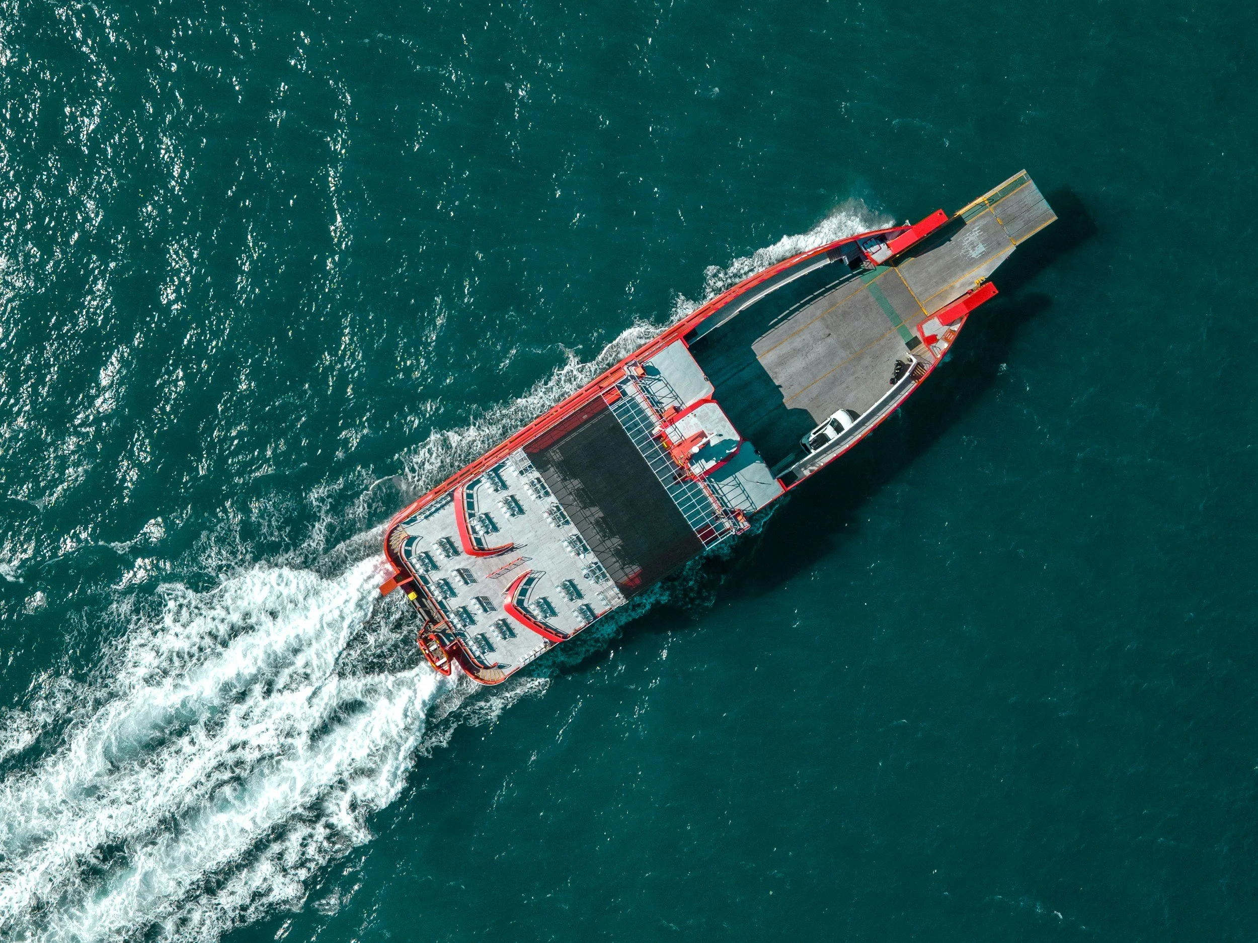 Aerial view of a red and black cargo ship sailing through turquoise waters, creating white wakes behind it.
