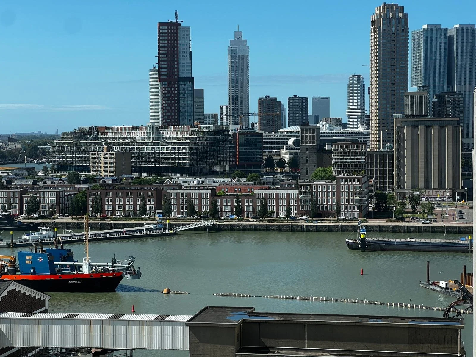 View of the Port of Rotterdam with tall modern buildings and a body of water in the foreground, including ships docked at the port.
